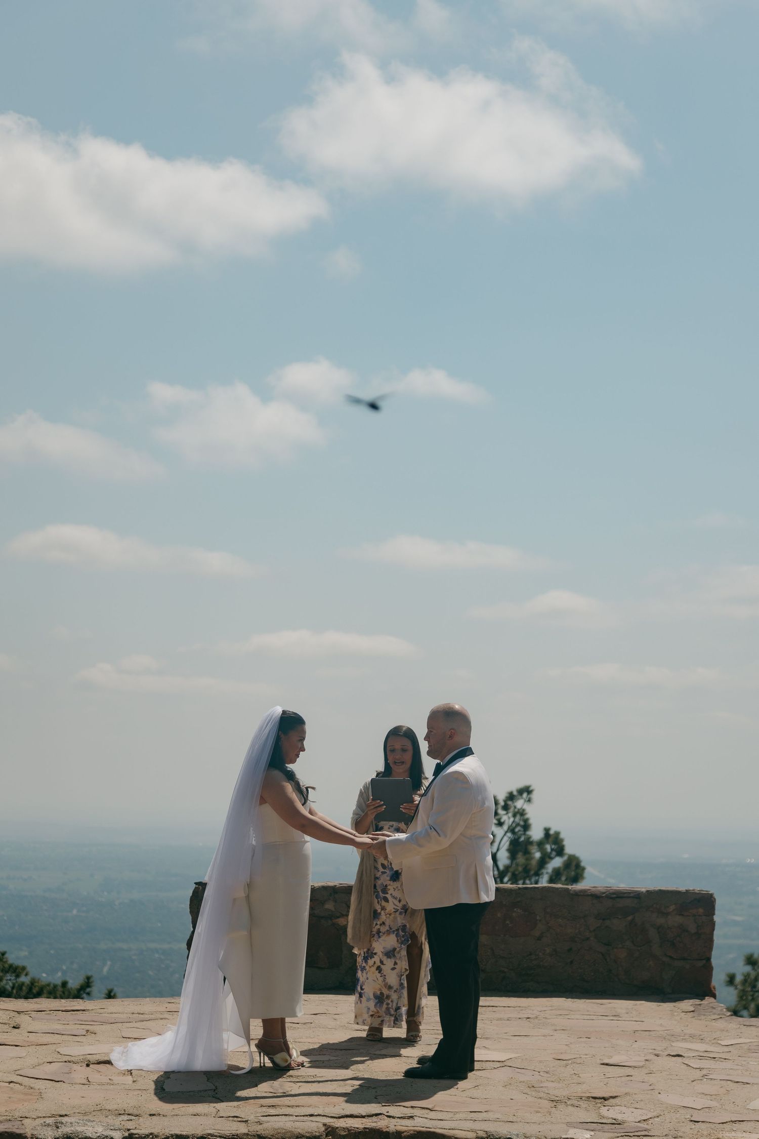 Bride and groom exchange vows outdoors on a stone terrace with a scenic panoramic view under a partly cloudy sky.