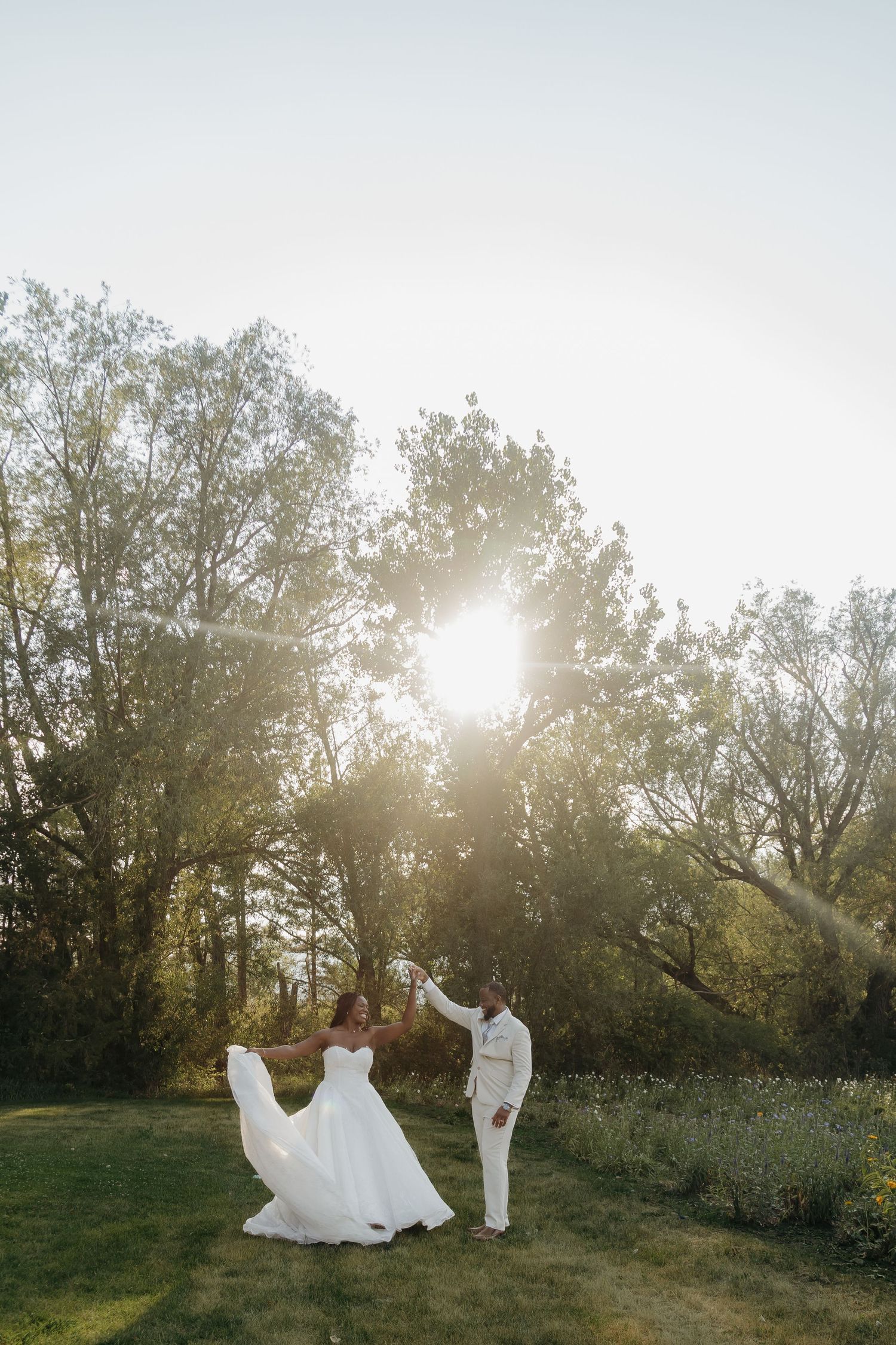 Bride and groom dancing outdoors at golden hour, surrounded by lush green trees with sunlight glowing behind them.