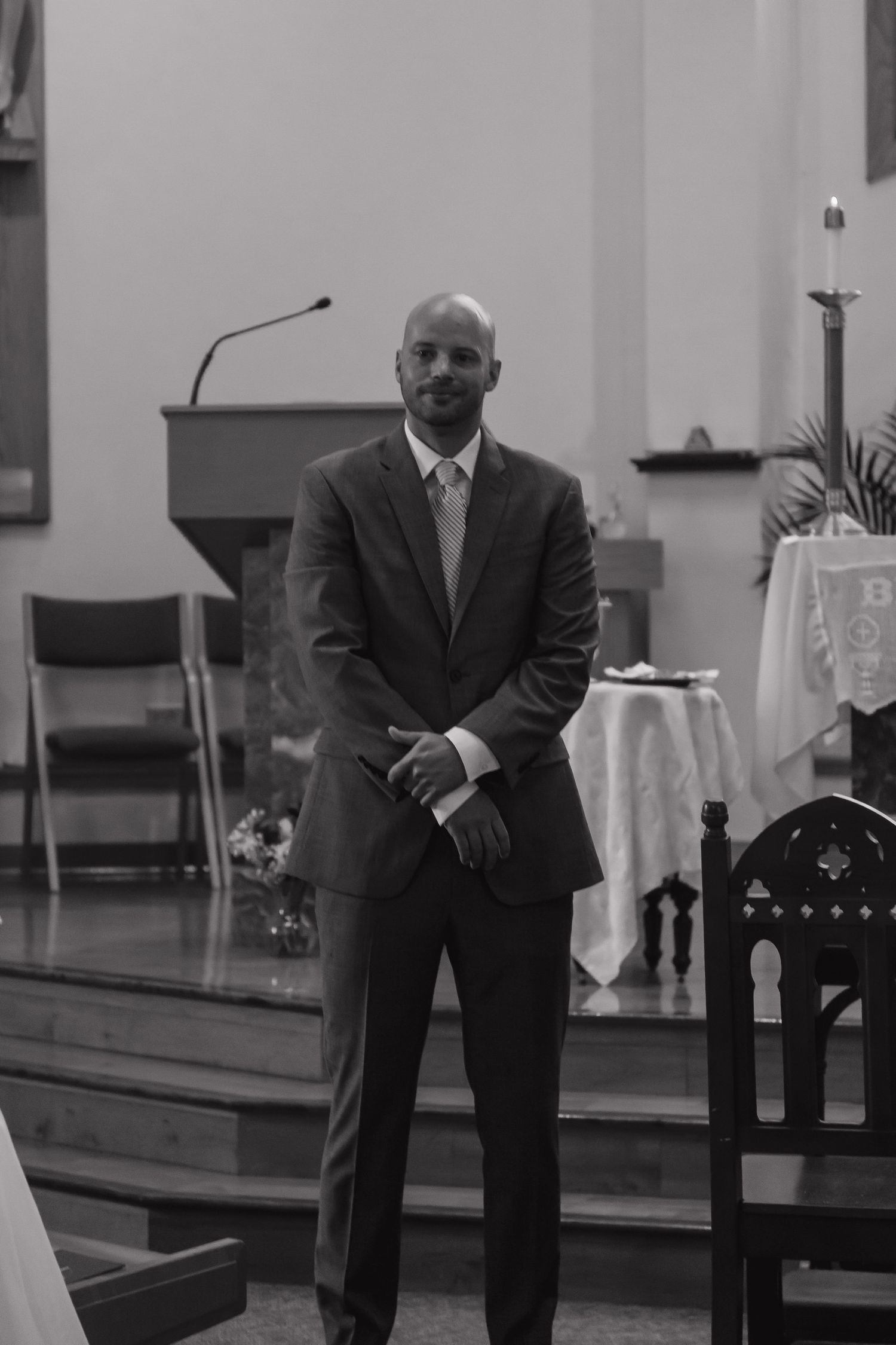 Groom standing at church altar in gray suit, smiling confidently before wedding ceremony, black and white photo.
