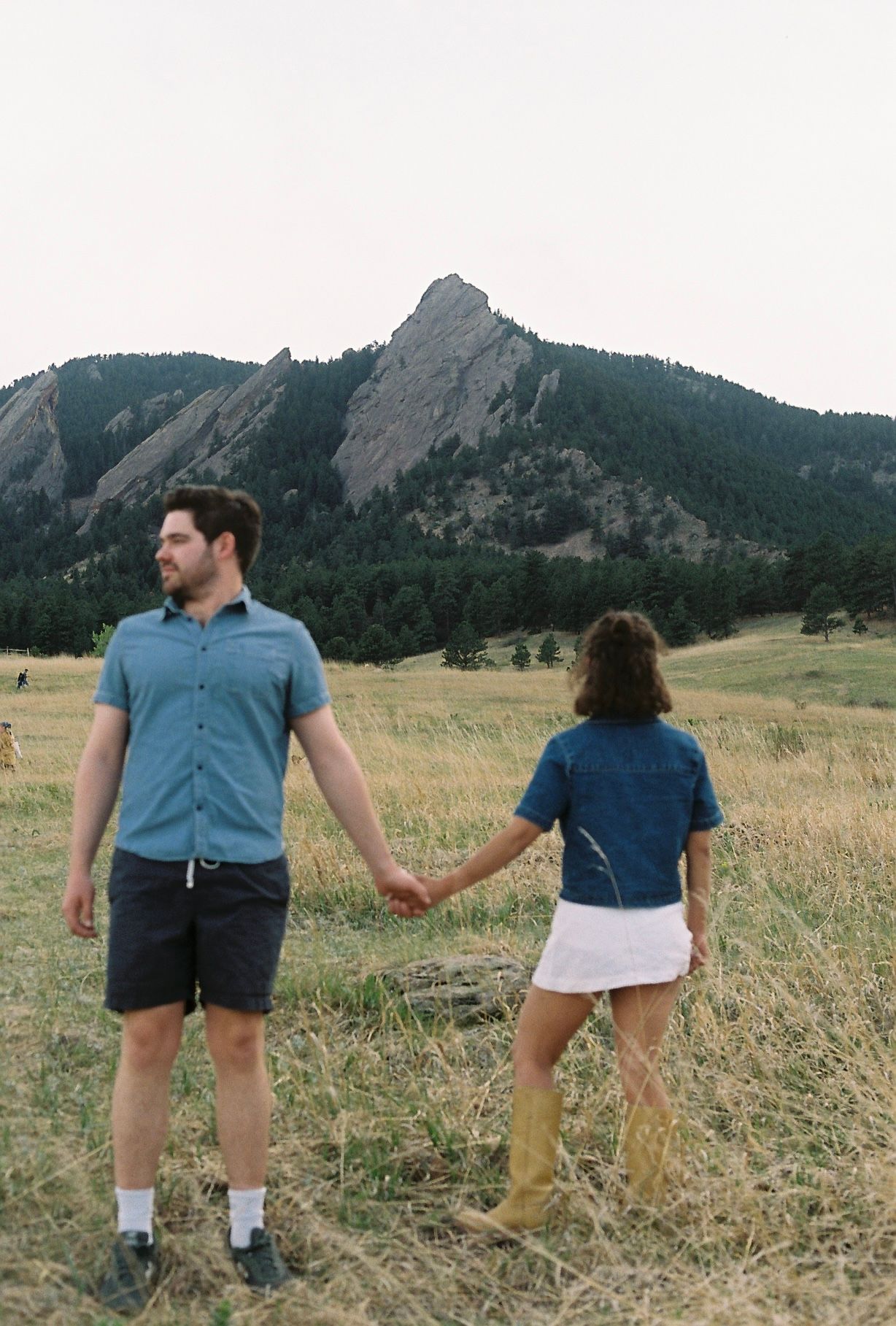A couple holds hands in a grassy field with the iconic Flatirons rock formations in Boulder, Colorado behind them.