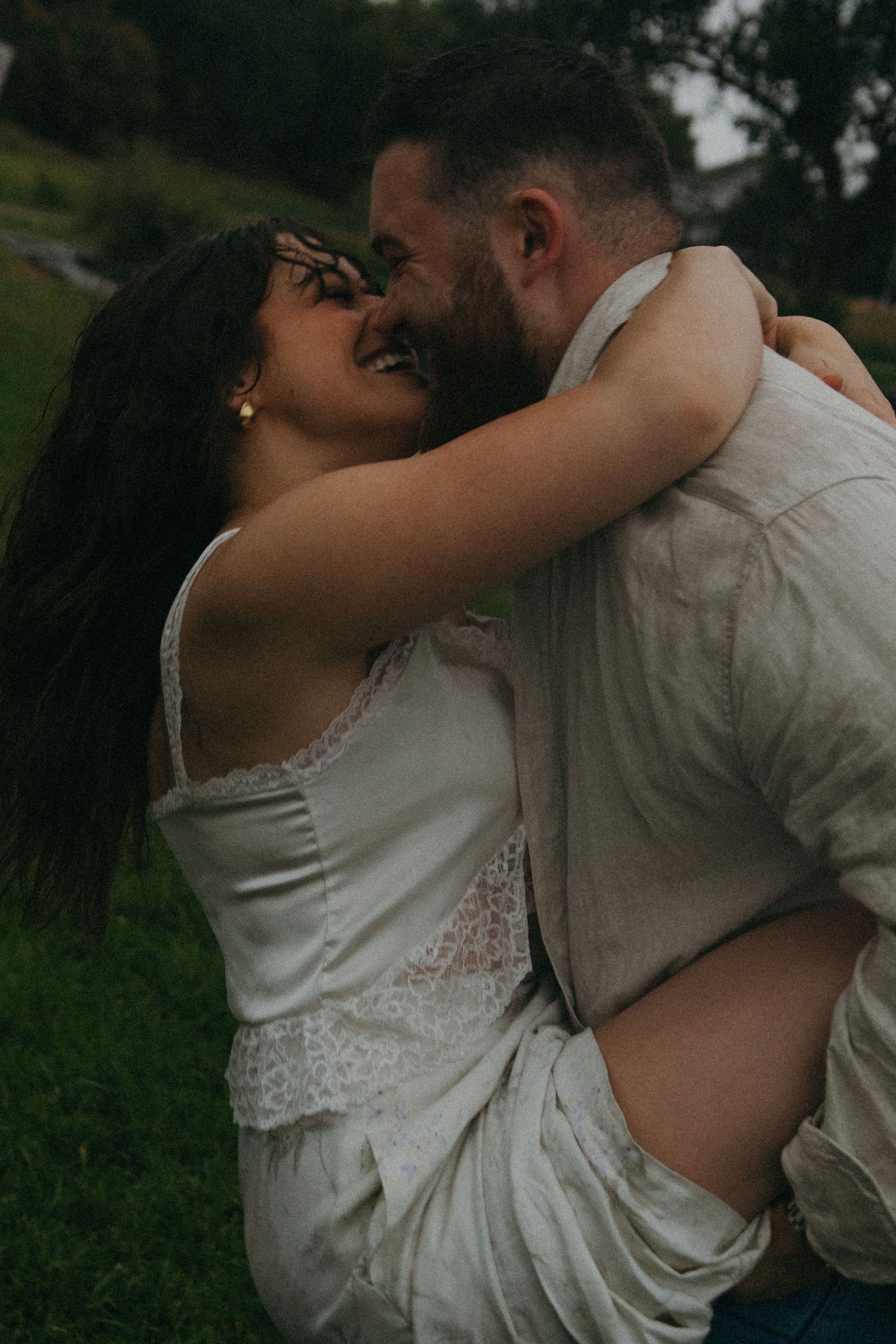 Couple sharing a joyful romantic moment outdoors, woman in white lace dress laughing as man lifts her in green field.