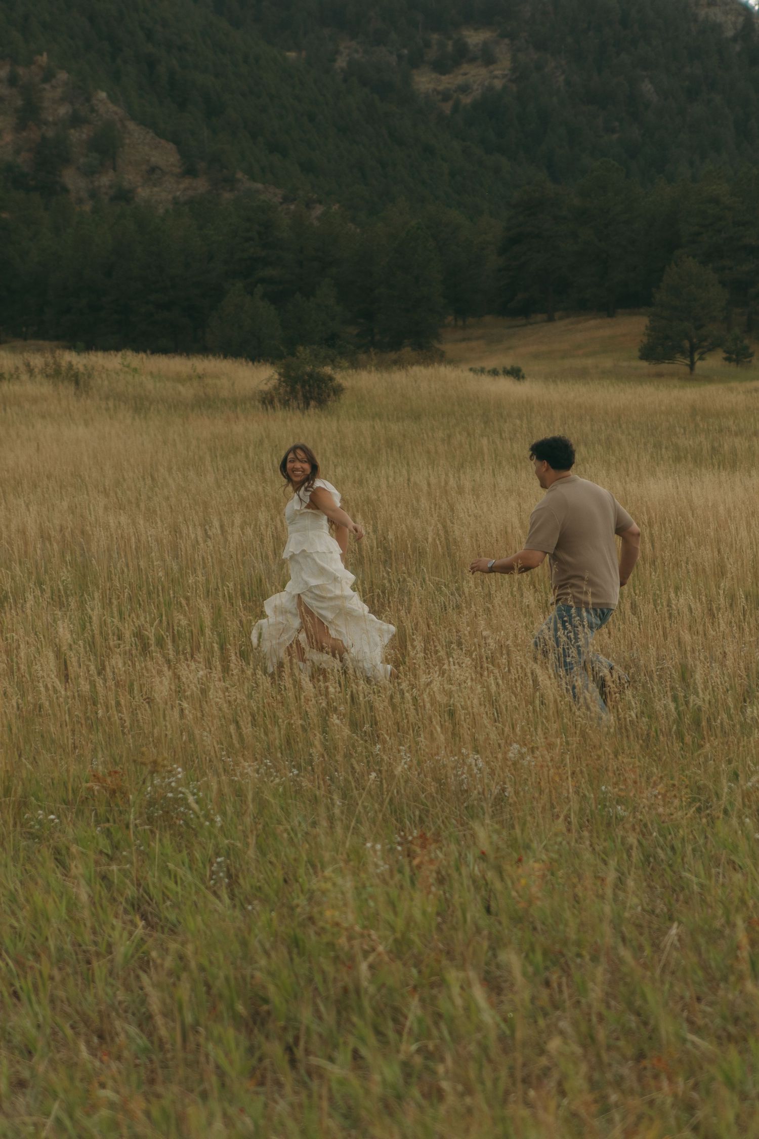Couple running through golden meadow with mountain forest backdrop during outdoor engagement photo session.