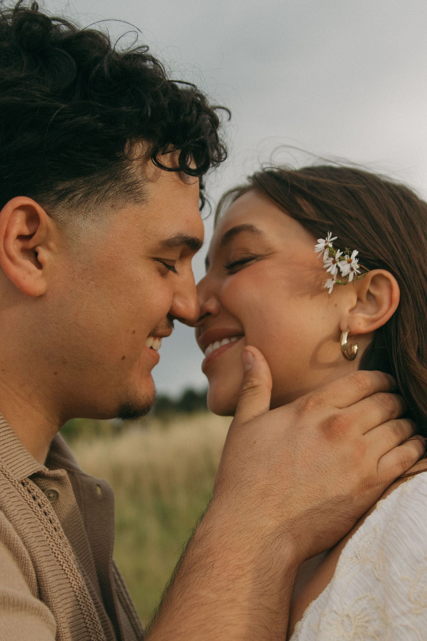 Romantic couple almost kissing in field, woman with flowers in hair, man gently holding her face.