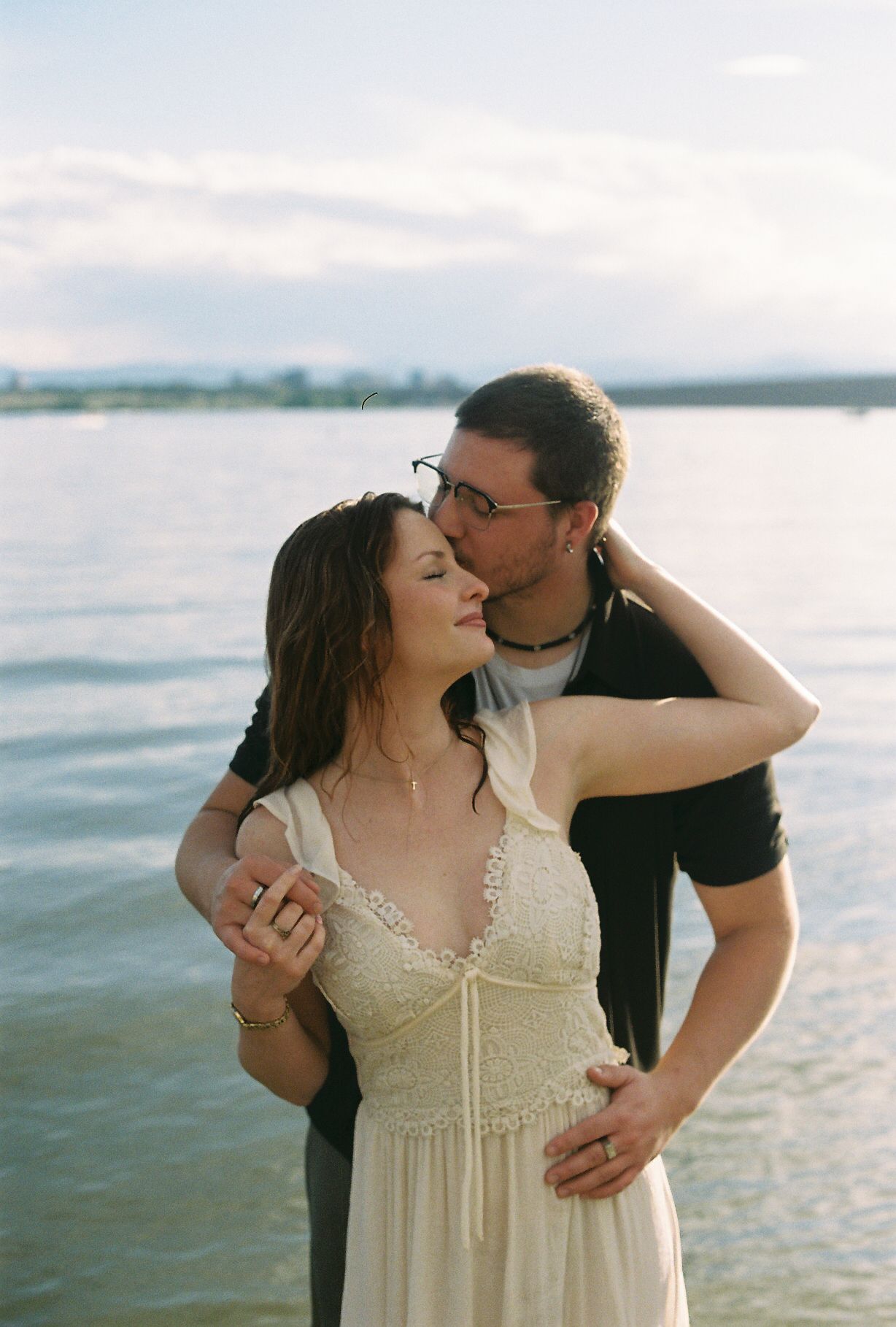 Man kissing woman on cheek while embracing her by a lake in soft natural light.
