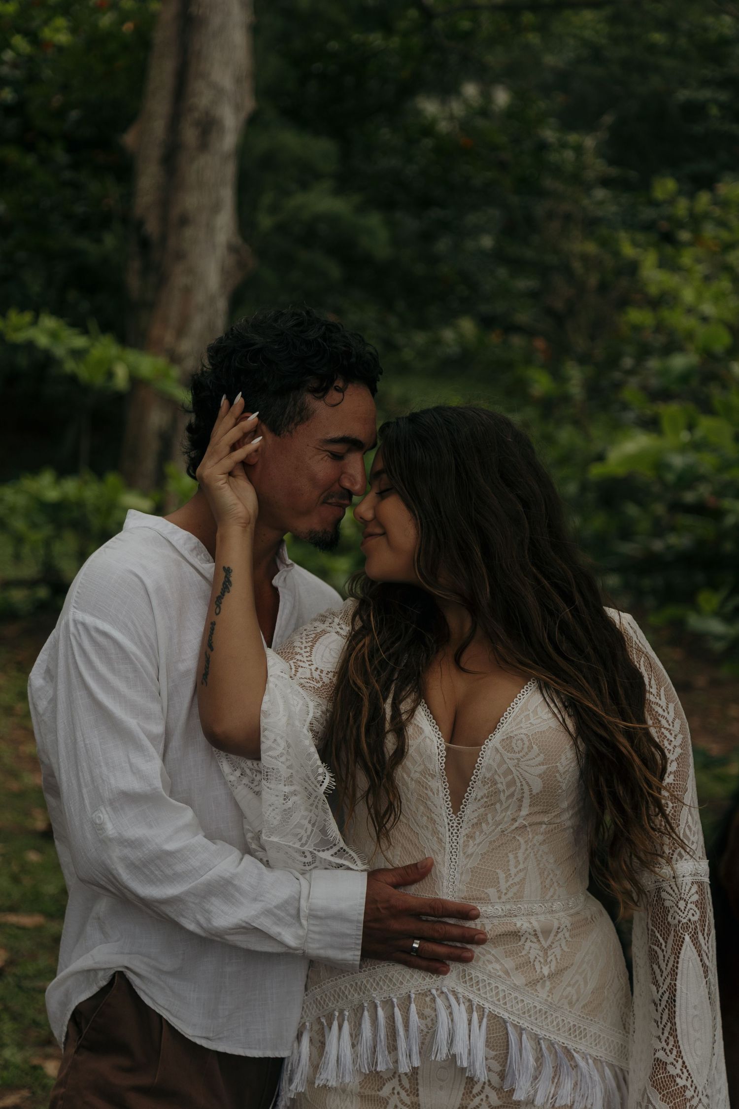 Couple almost kissing in lush green forest, bride in boho lace dress holds grooms face tenderly.