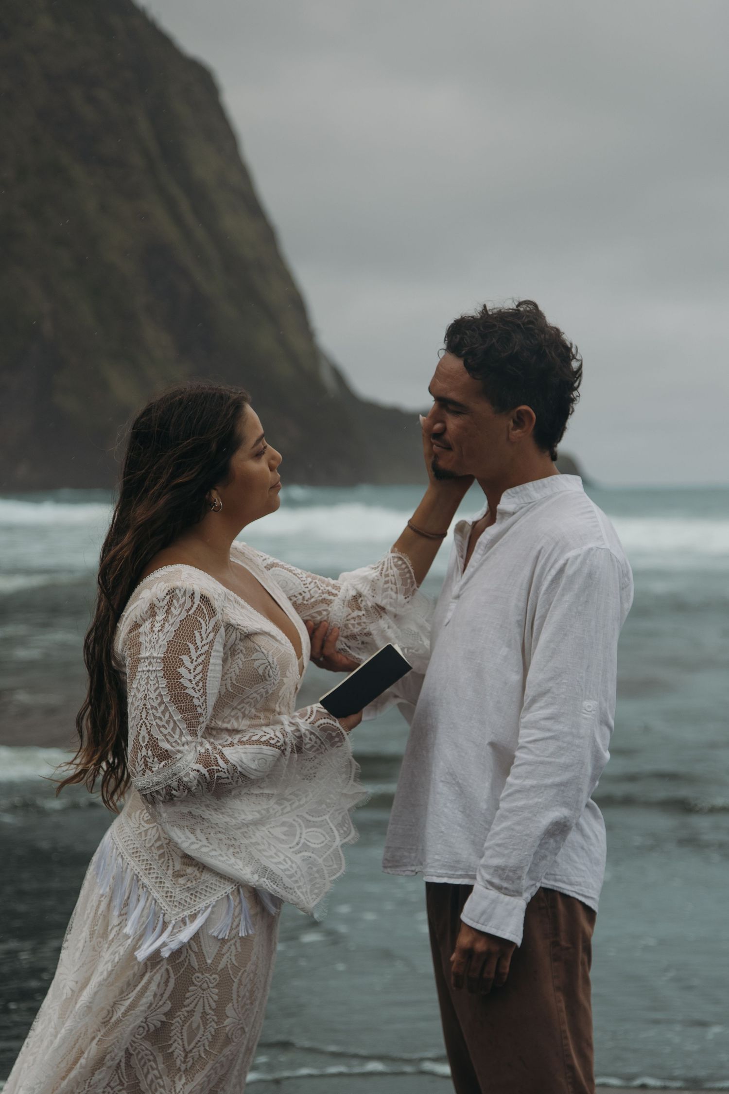 Bride tenderly touches groom's face during beach elopement ceremony with dramatic ocean cliffs behind.