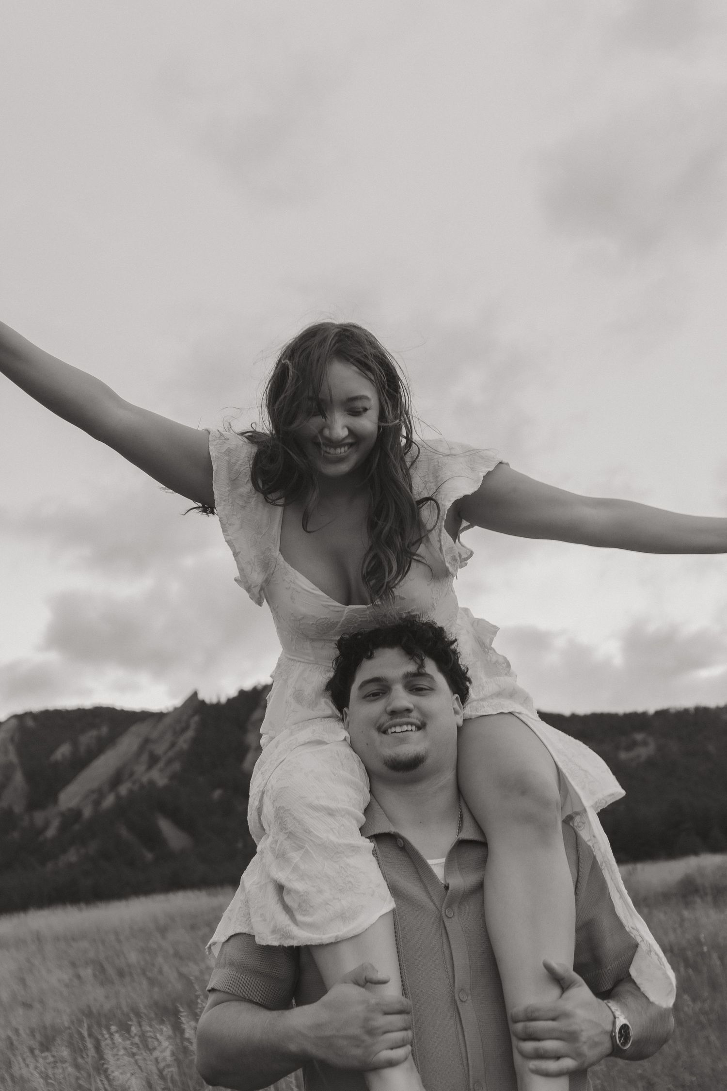 Black and white photo of woman sitting on mans shoulders, arms spread wide, laughing outdoors.