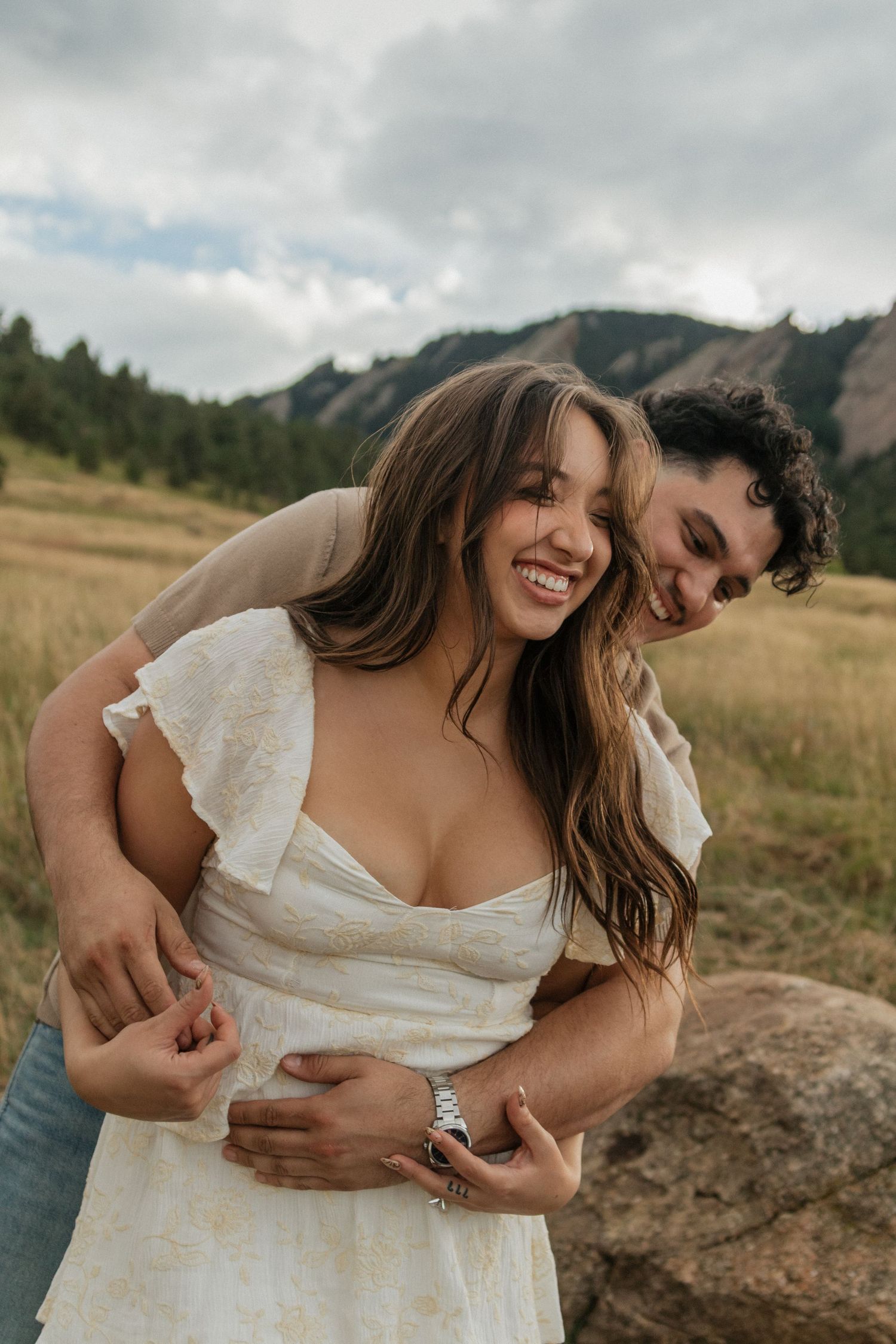 Laughing woman in white dress being embraced by her partner in a scenic mountain meadow engagement photo.