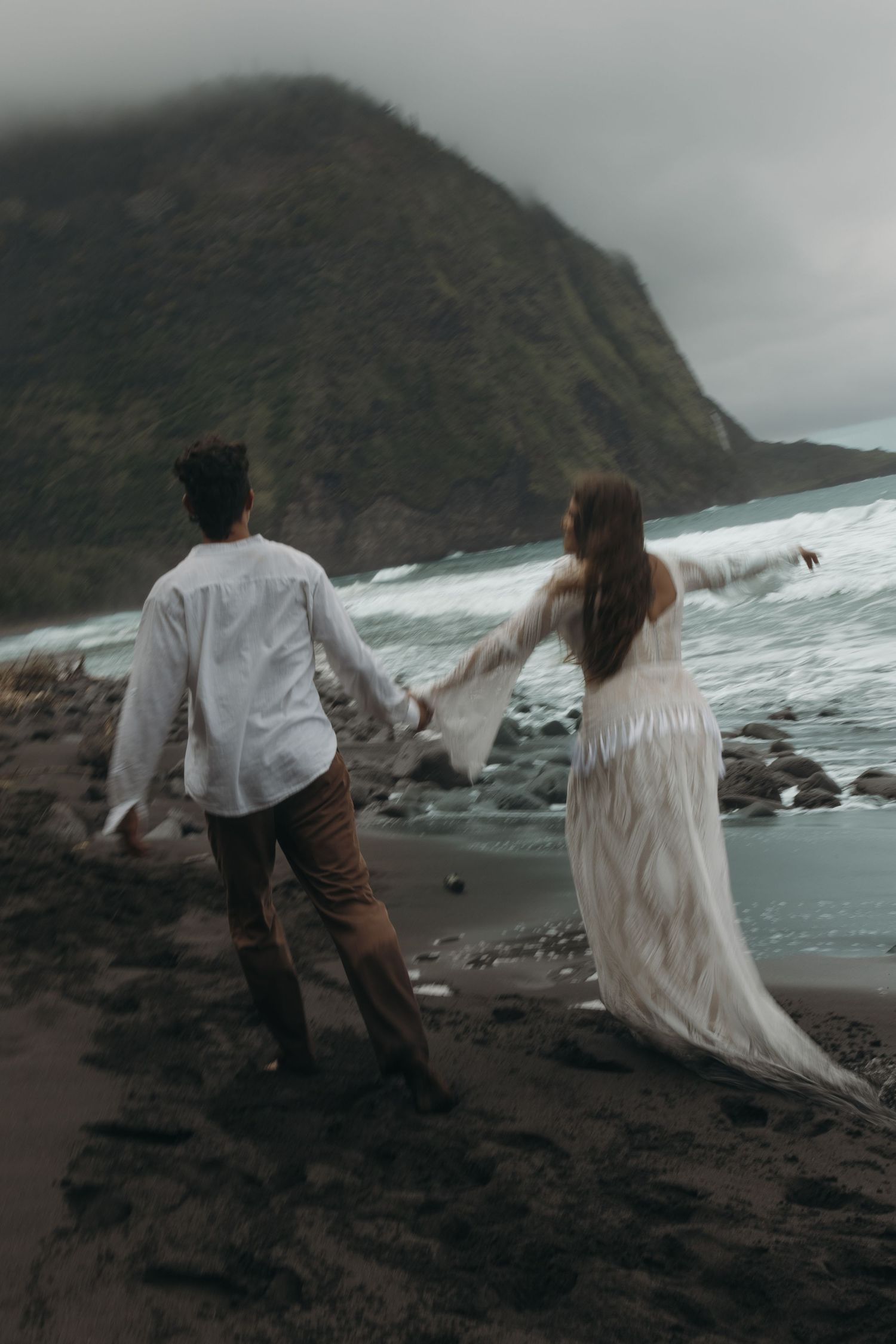 Couple walks hand-in-hand toward crashing waves on black sand beach with misty green cliffs.