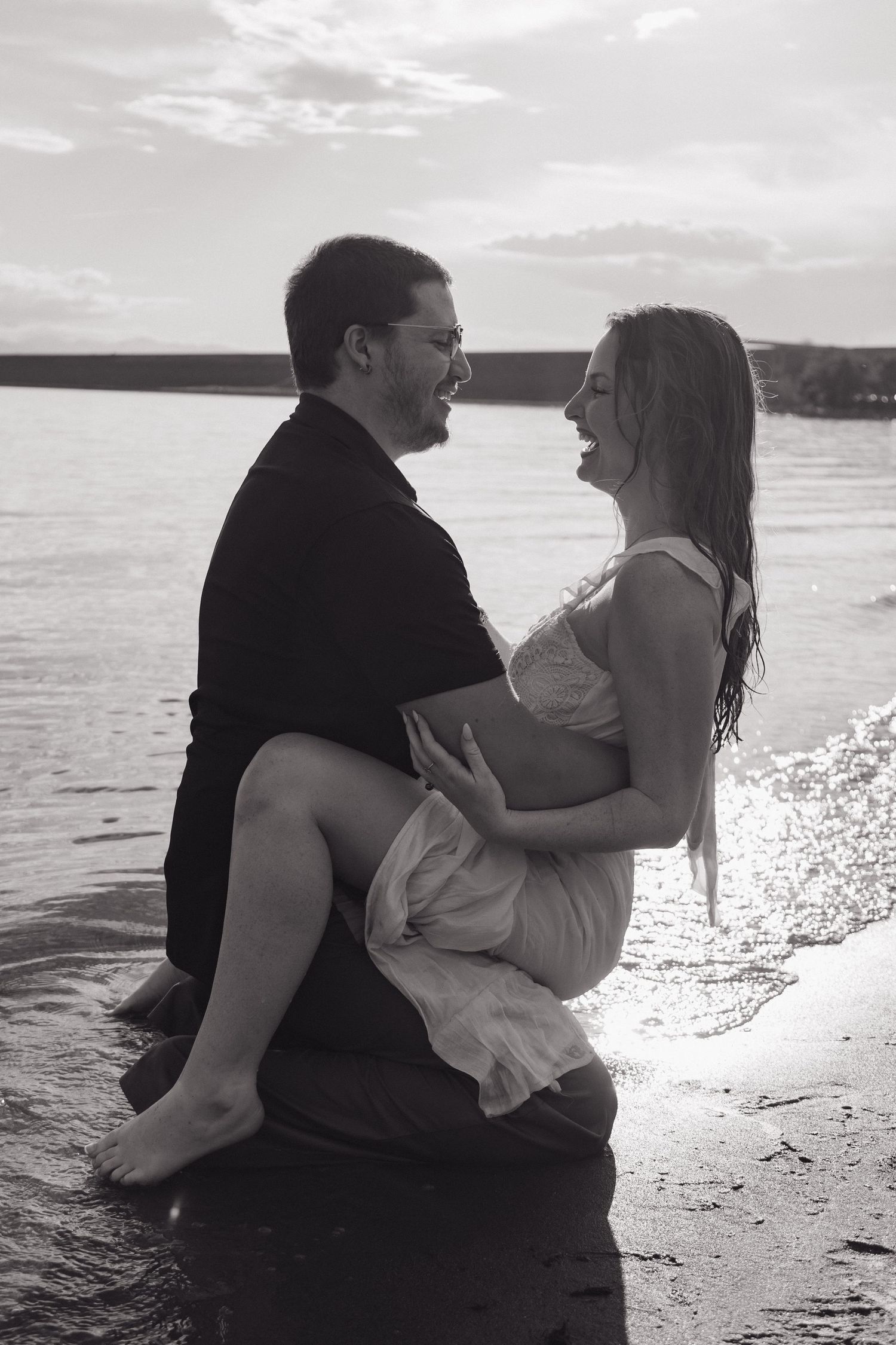 Black and white photo of laughing couple embracing in shallow water during beach photo session.