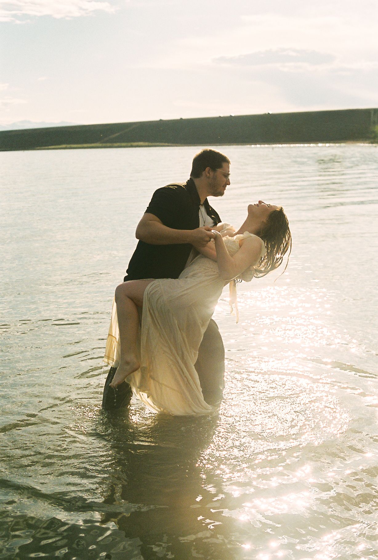 Man dips woman in white dress dramatically in sunlit lake during romantic couples photo session.