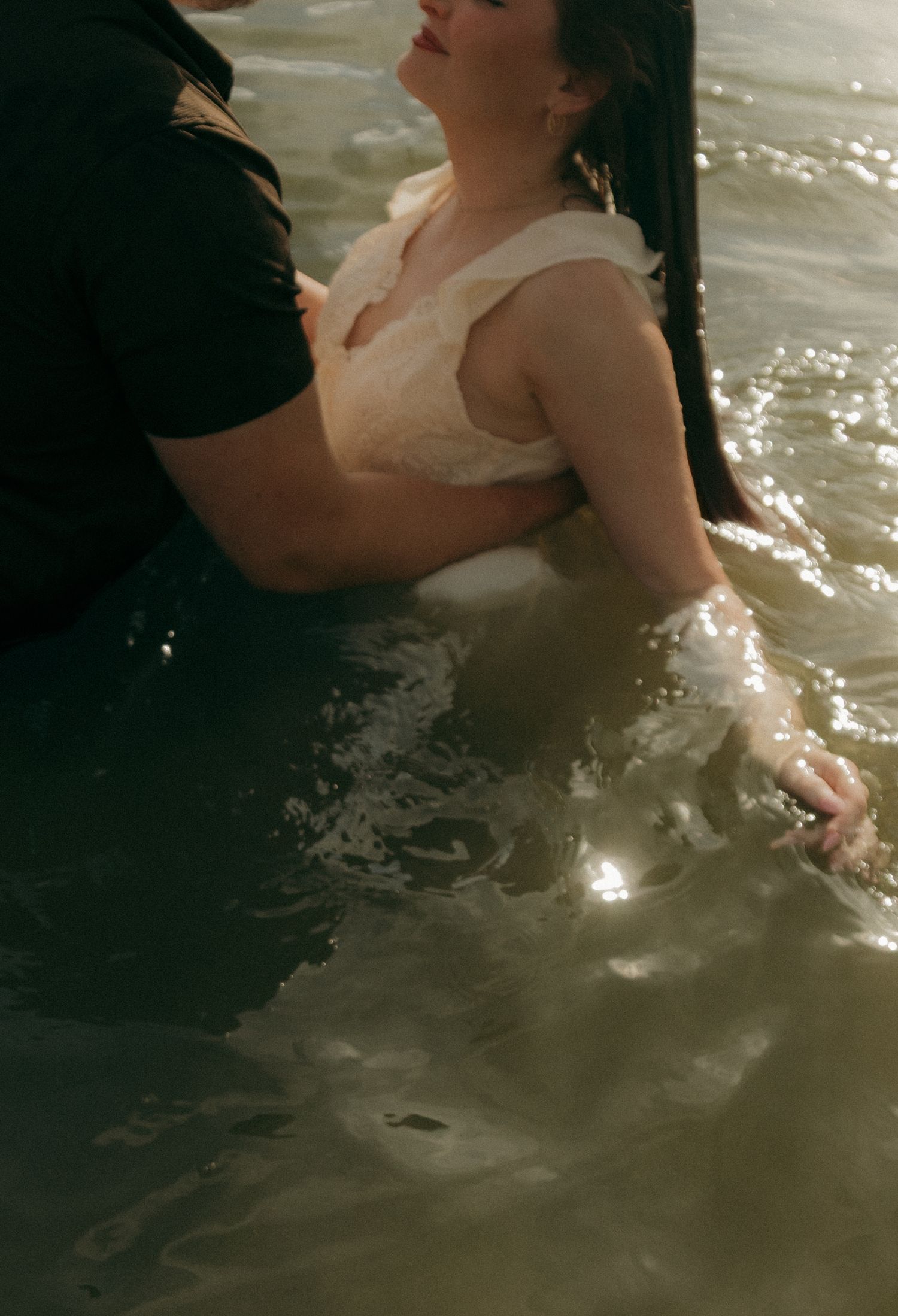 Close-up of couple submerged in shimmering water, woman in white lace top embraced by partner in black.