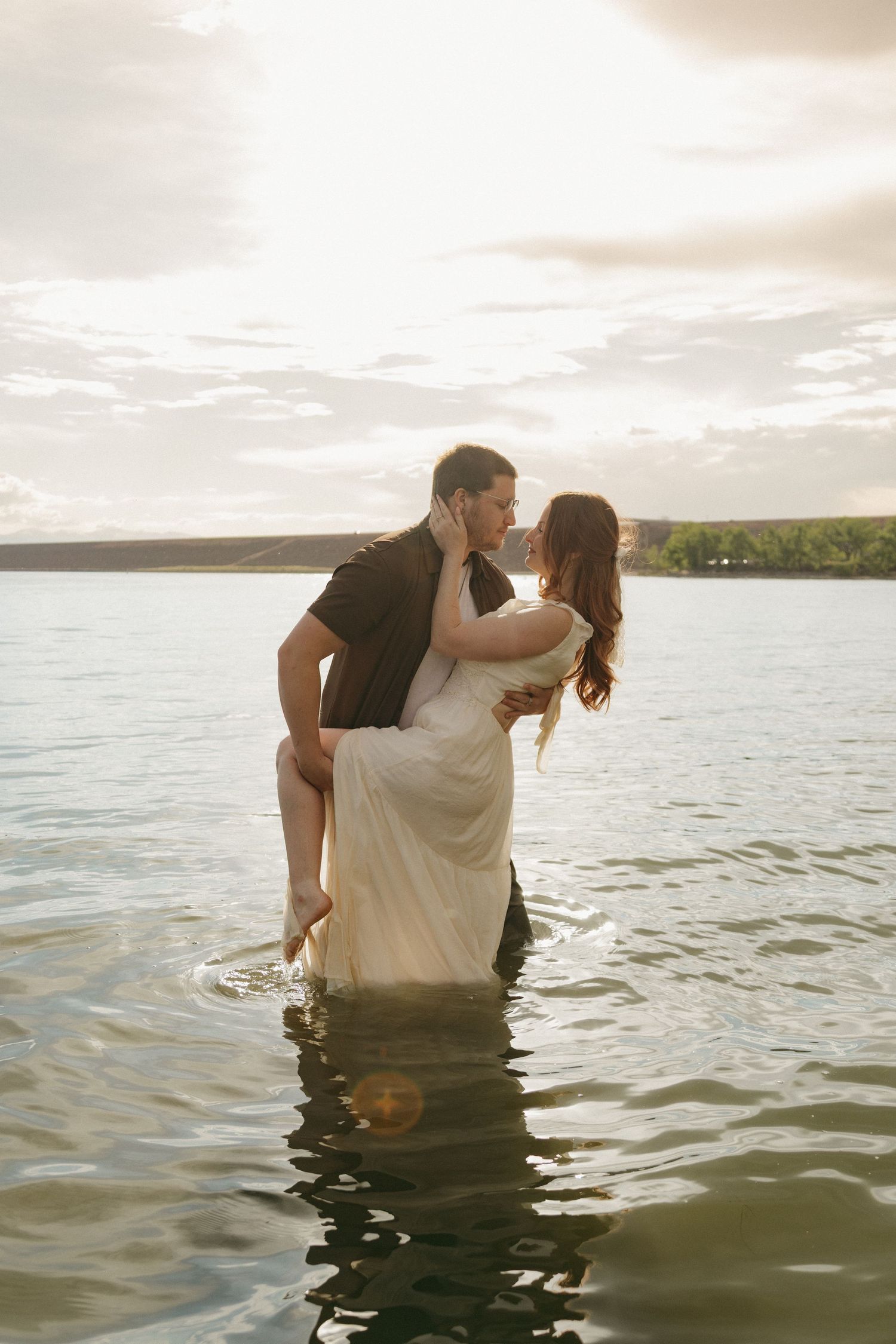 Romantic couple embracing and kissing while standing in a lake at golden sunset hour.