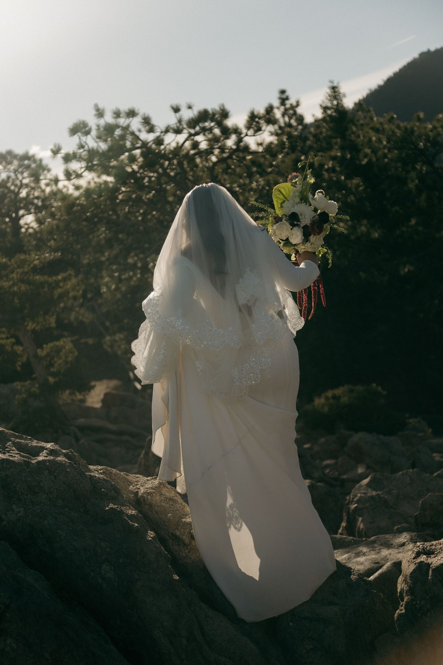 Bride in white gown and veil holds bouquet of anemones while standing on rocky mountain terrain at sunset.
