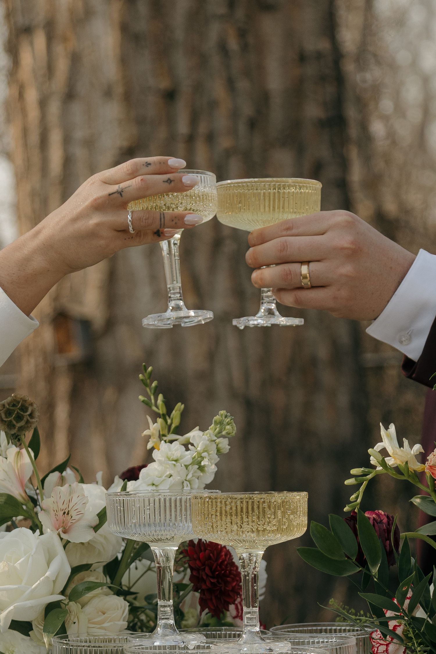 Bride and groom toasting with elegant champagne coupe glasses over floral champagne tower at outdoor wedding.