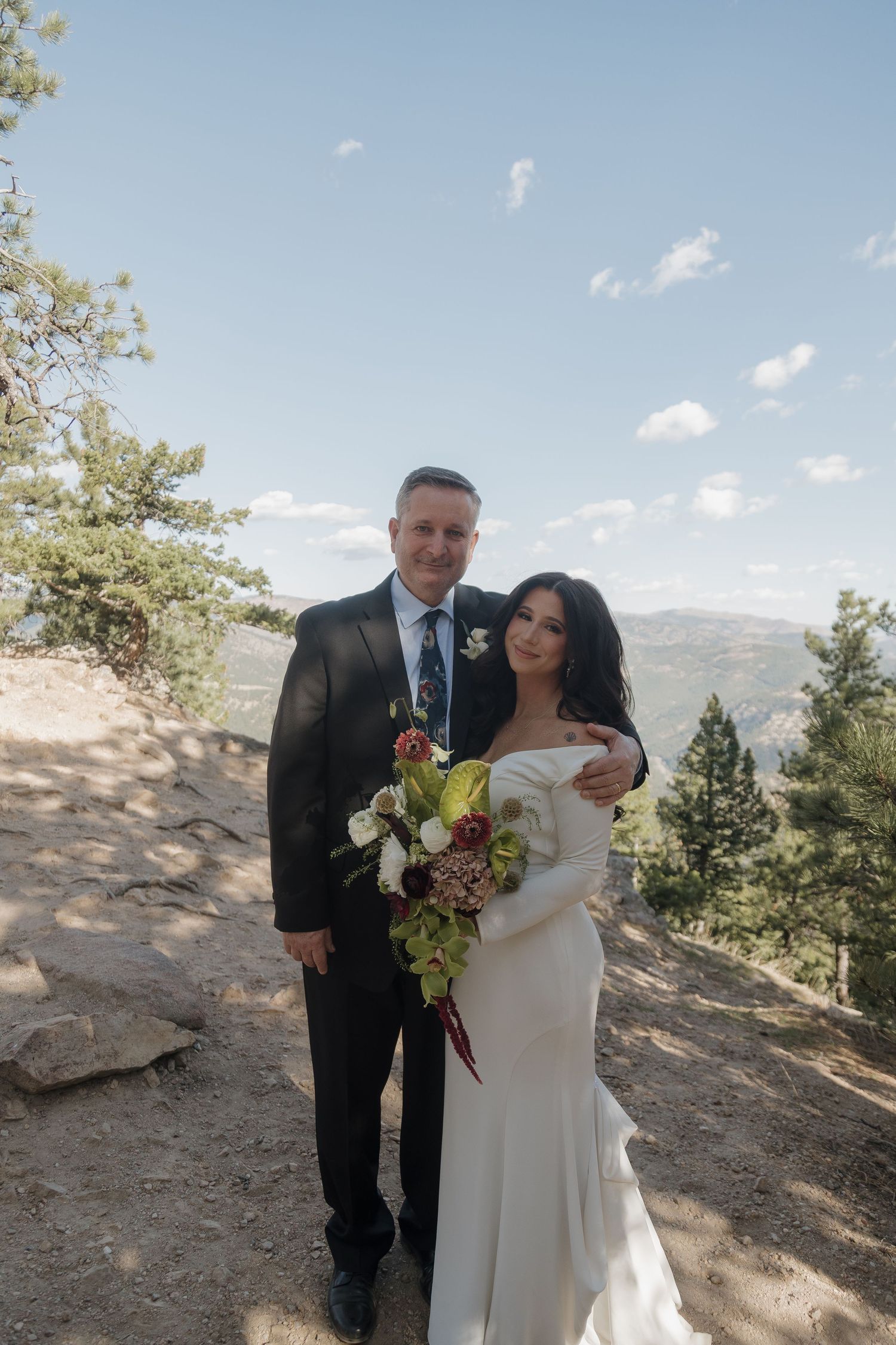 Bride and groom posing together on a mountain overlook, she holds a bouquet in a white off-shoulder gown.