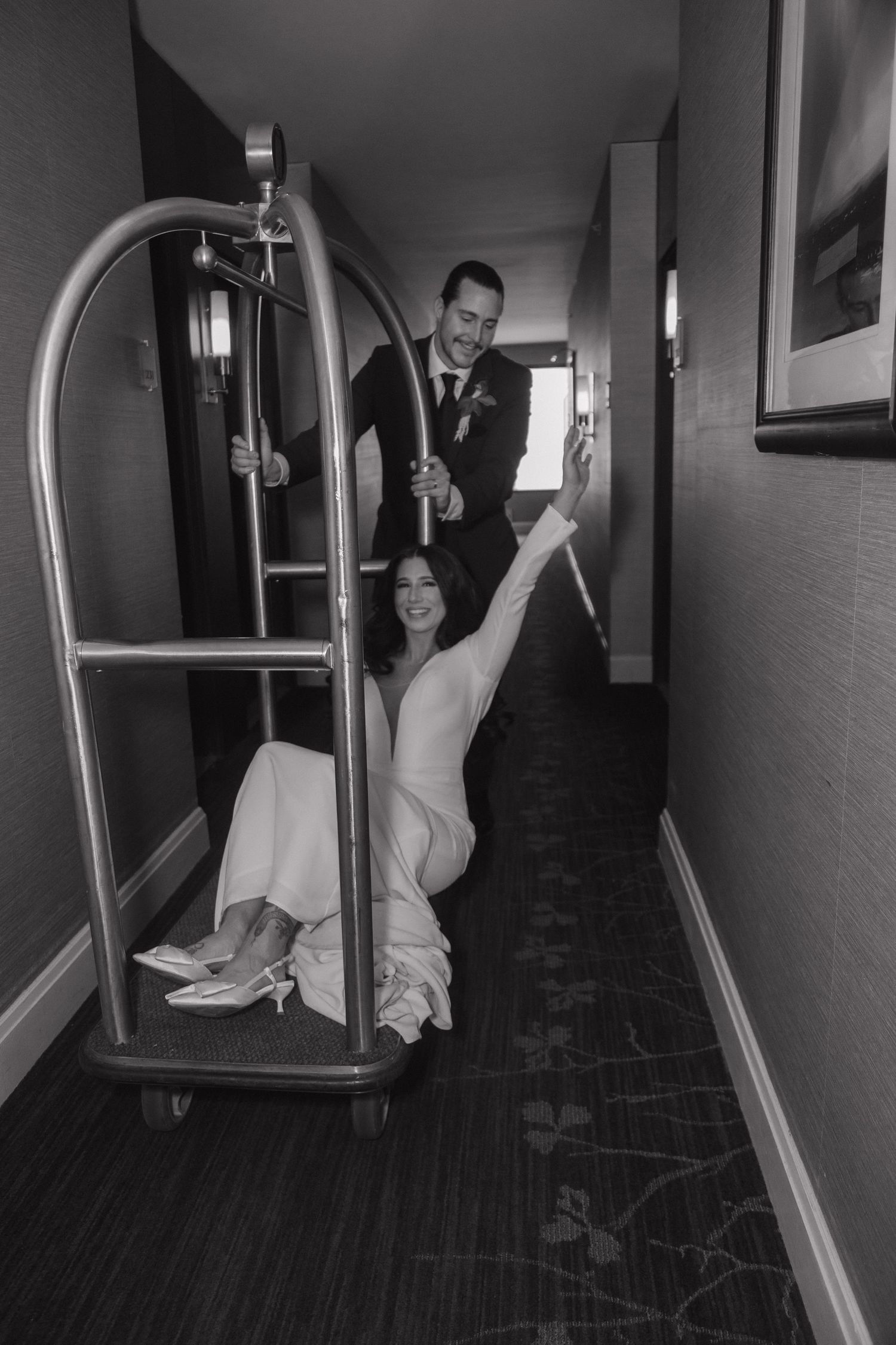 Playful black and white photo of bride riding hotel luggage cart pushed by groom.