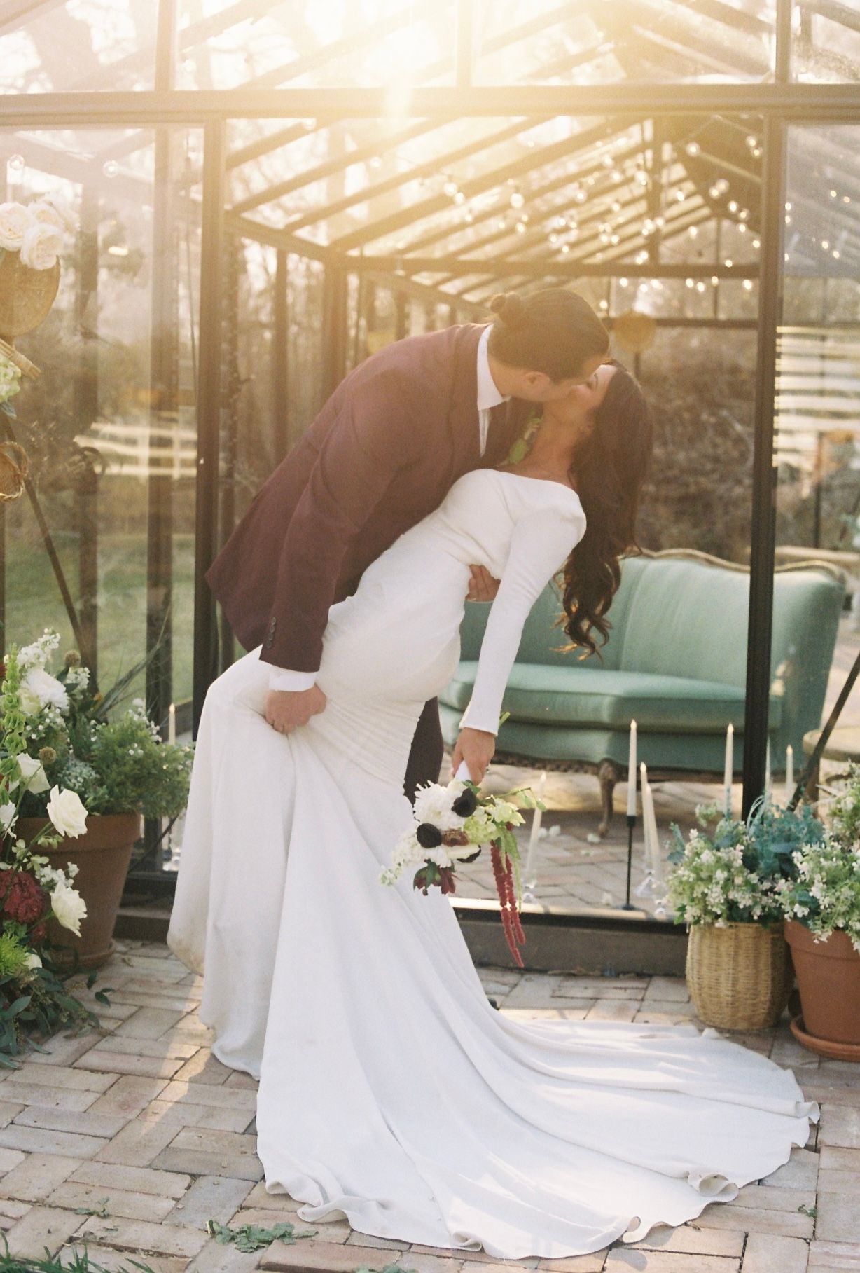 Groom dips bride for a kiss in front of a sunlit greenhouse surrounded by lush greenery.