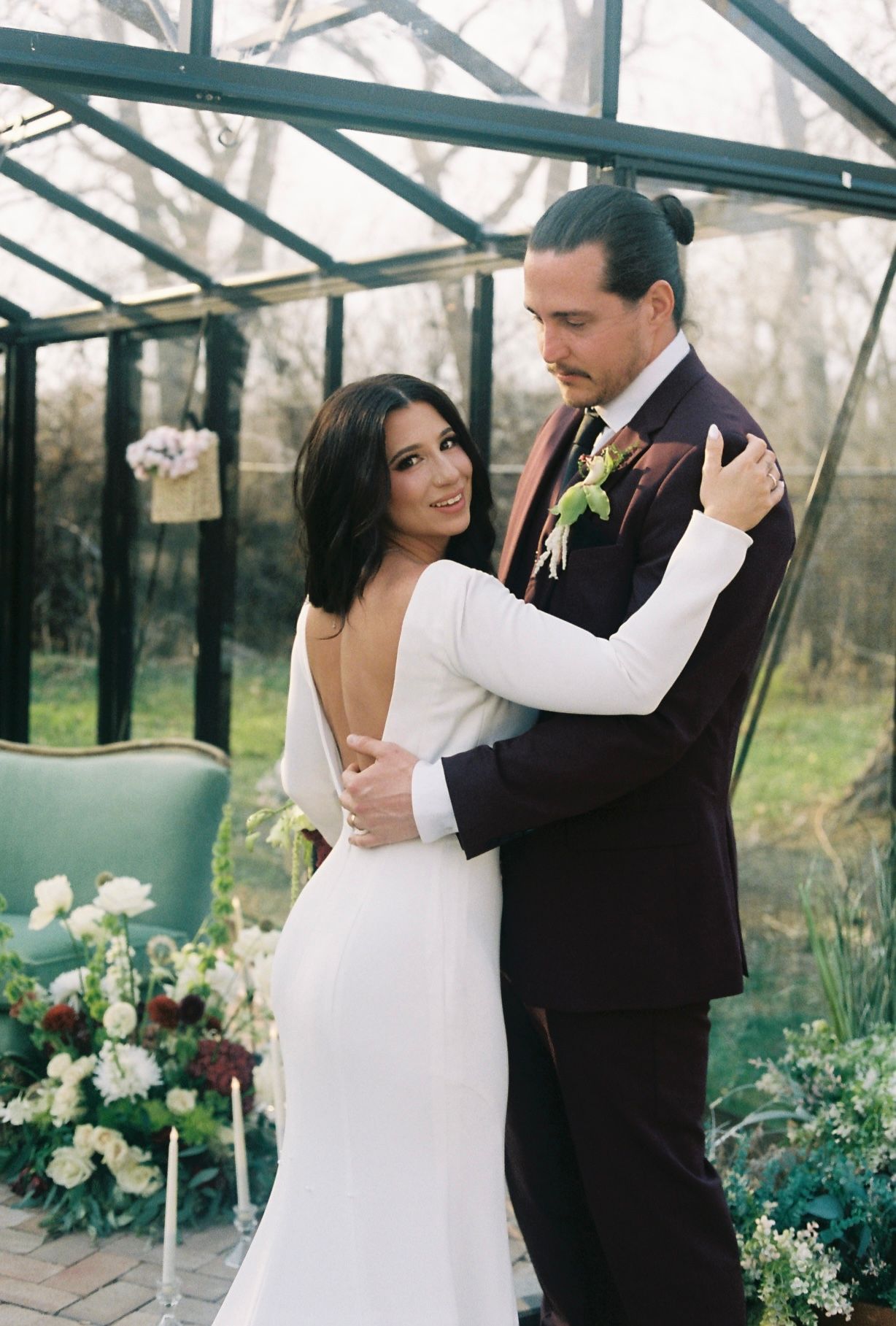 Bride in backless white gown embraces groom in burgundy suit before a glass greenhouse with floral decor.