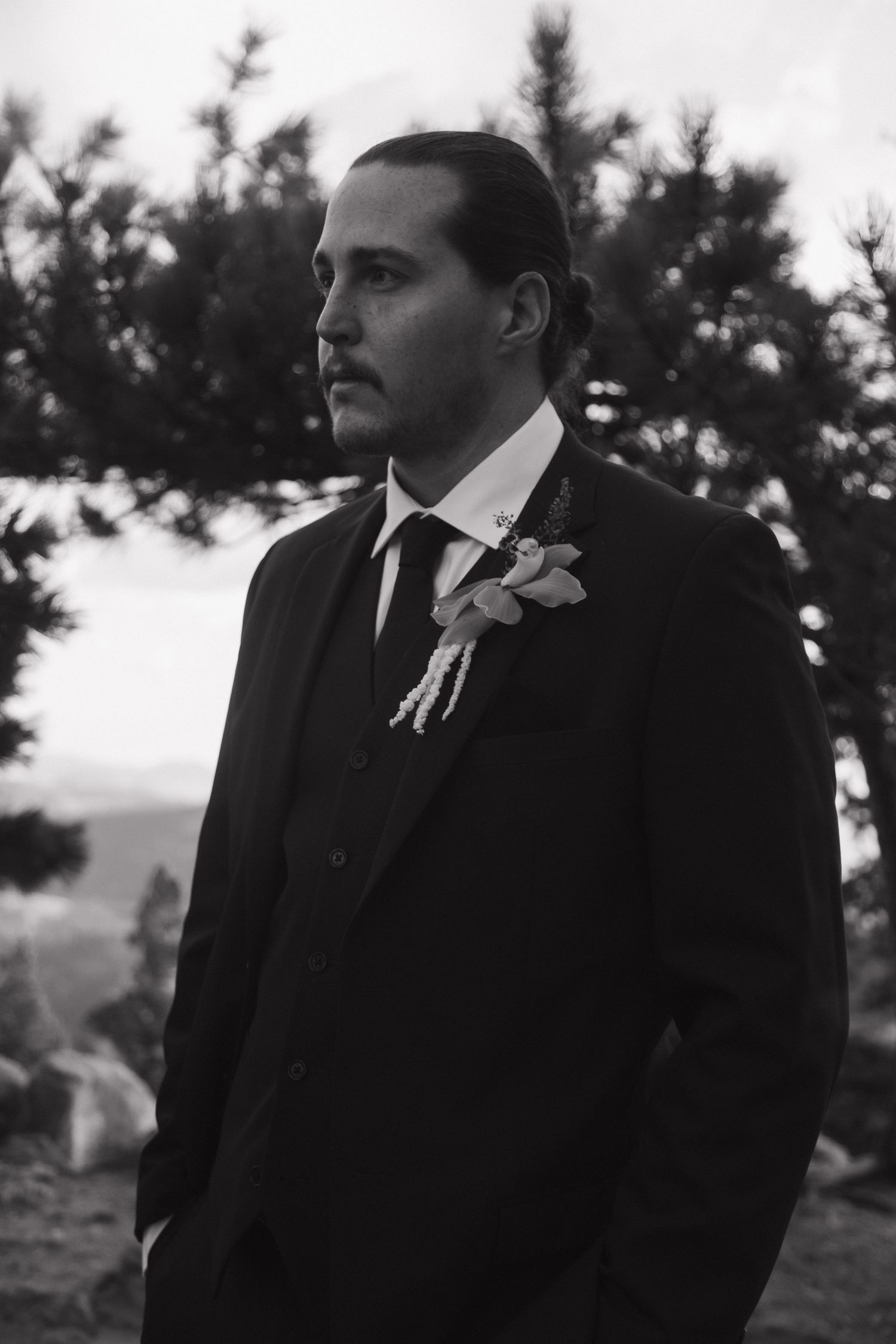 Black and white portrait of groom in dark suit with boutonniere among pine trees.