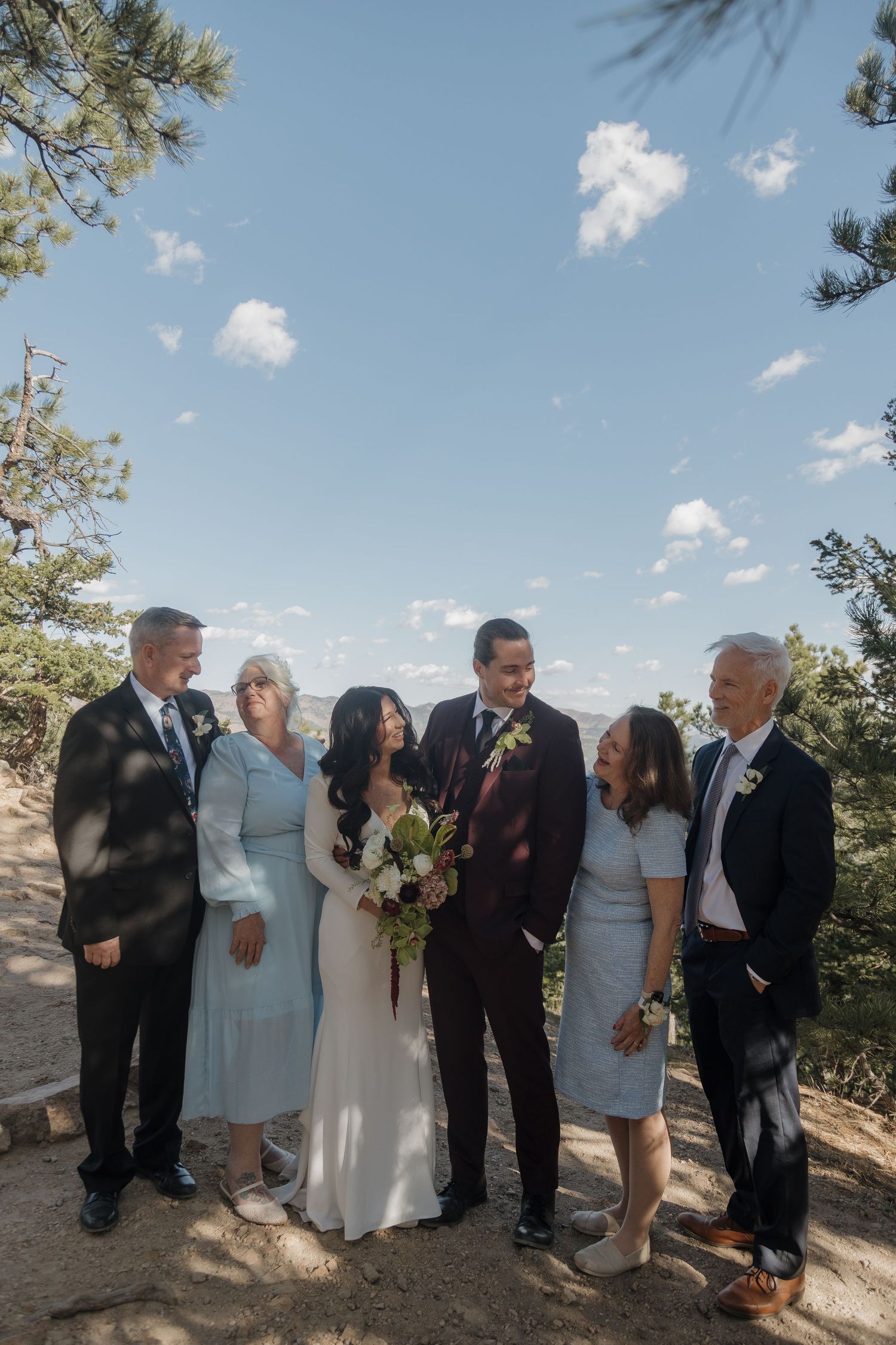 Bride and groom pose with four family members outdoors under a blue sky surrounded by pine trees.