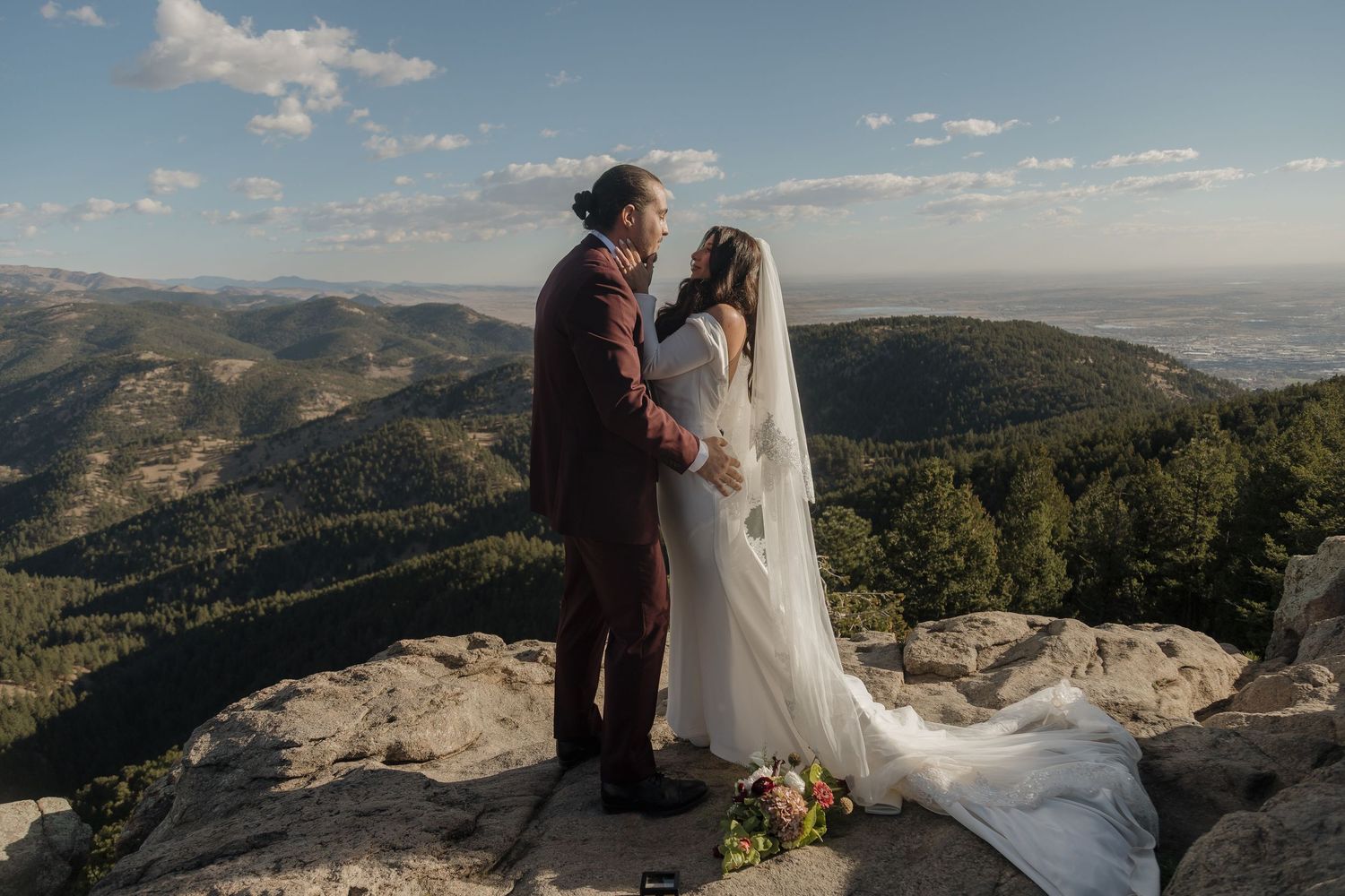 Couple in wedding attire sharing intimate moment on rocky mountain peak with stunning panoramic valley views.