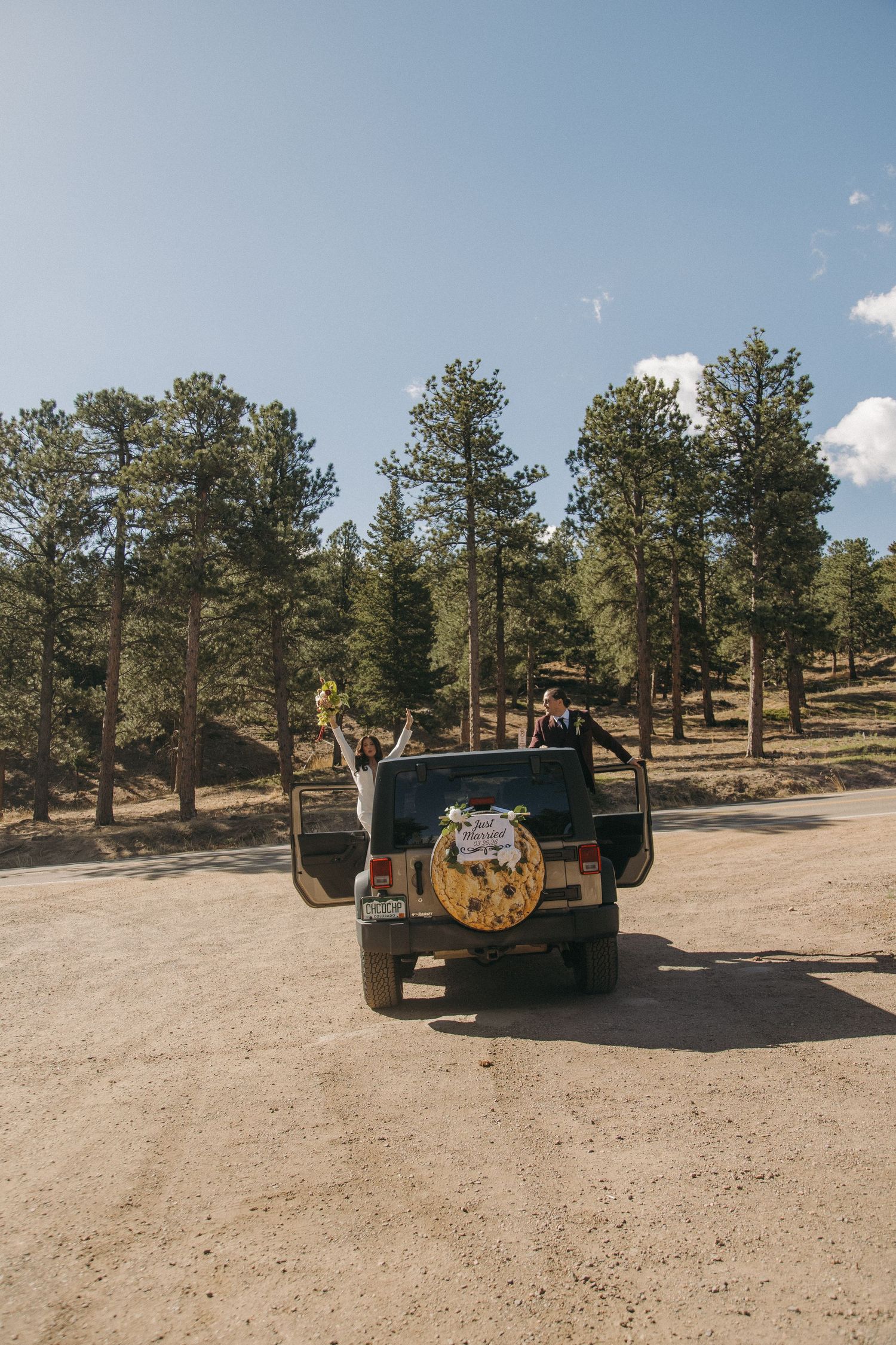Bride and groom celebrate beside a decorated Jeep on a dirt road surrounded by pine trees.