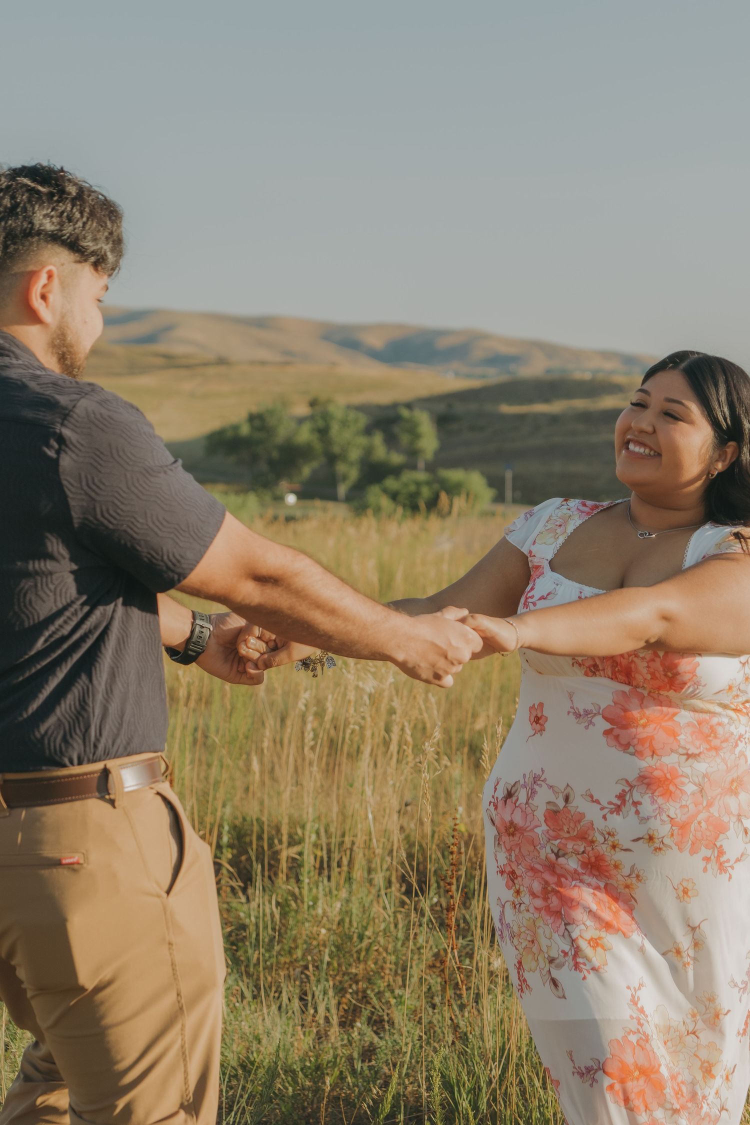 A couple holds hands and dances together in a scenic field with rolling hills in the background.