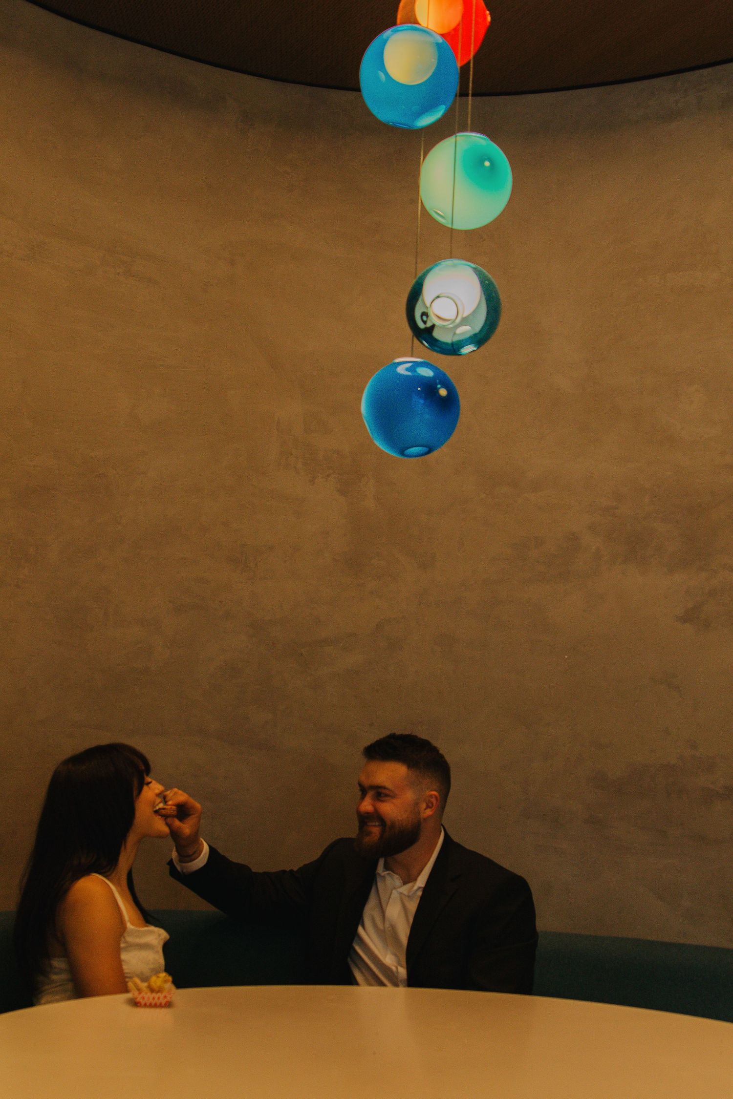 A couple sits at a table under colorful hanging pendant lights in a moody setting.