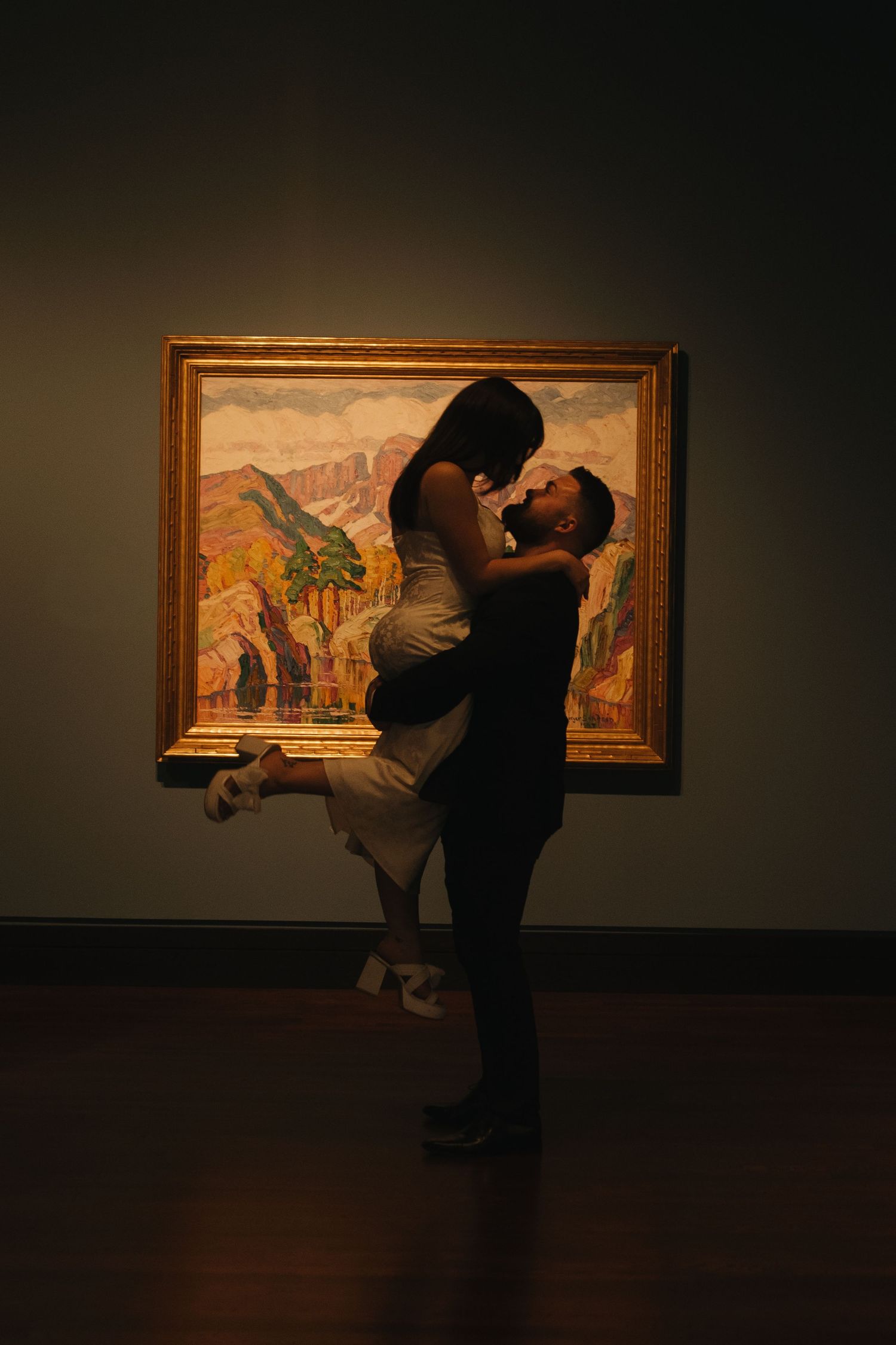 A couple shares tender kisses while viewing artwork in a dimly lit museum gallery.