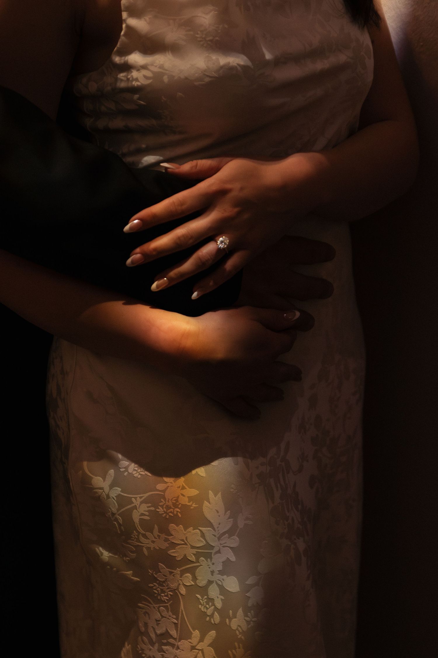 Close-up detail of hands against an elegantly textured white dress with intricate embroidery in moody lighting.