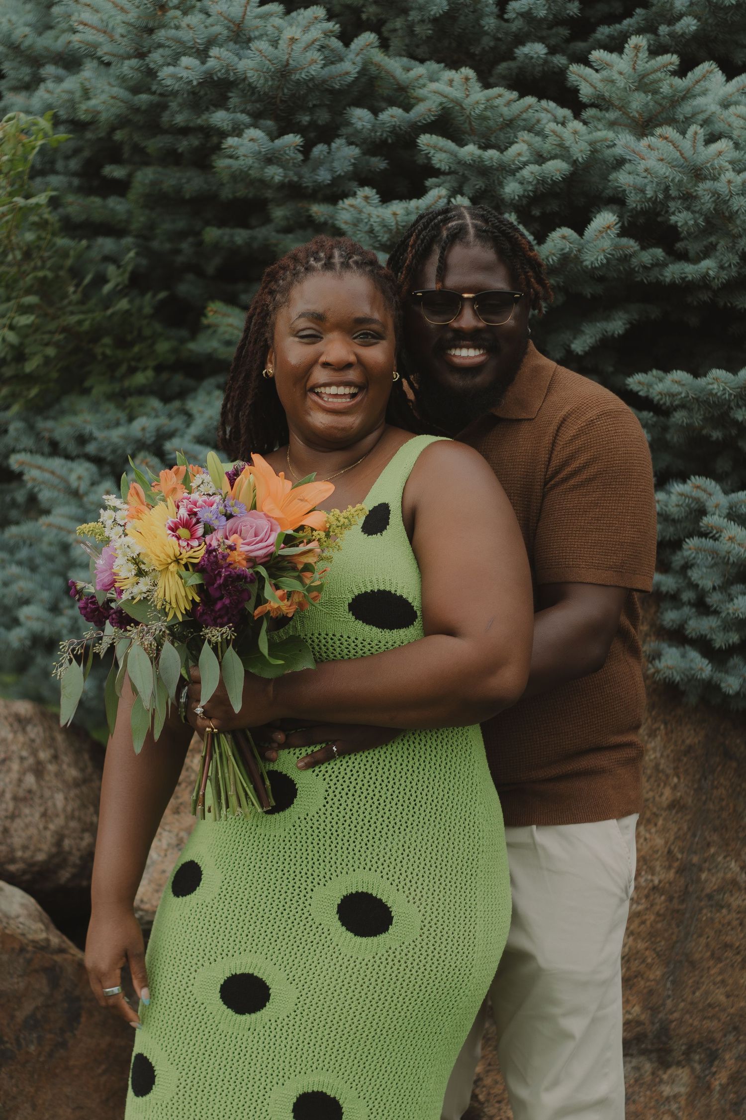 A loving couple poses with a vibrant bouquet against blue spruce trees in an outdoor setting.