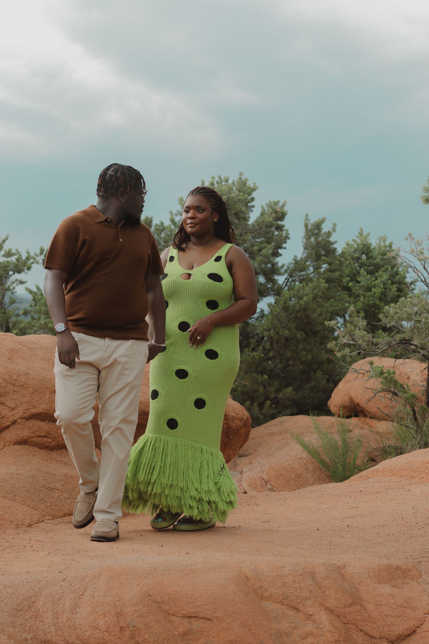 A romantic couple stands on red rocks at sunset, one wearing a bright lime green polka dot dress.