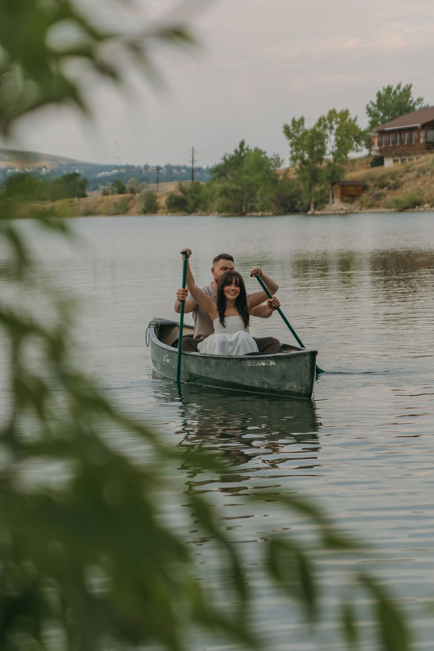 A couple rows a small boat across a tranquil lake surrounded by mountain scenery on a cloudy day.