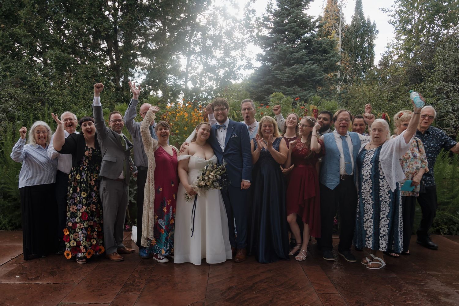 Wedding party group photo with bride in white gown and groom in navy suit, surrounded by cheering guests outdoors.