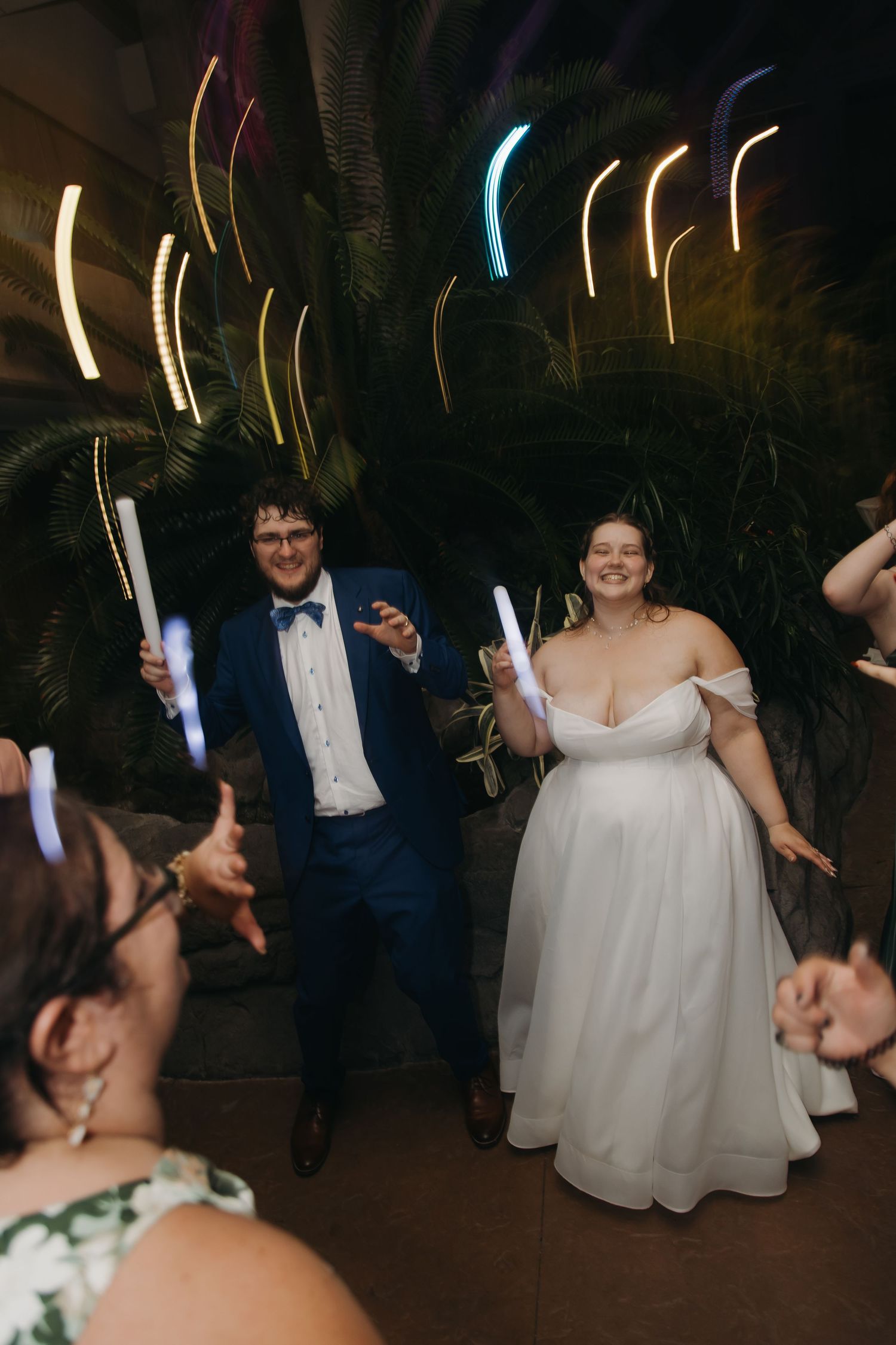 Bride and groom enter reception holding light wands surrounded by cheering guests and tropical decor.