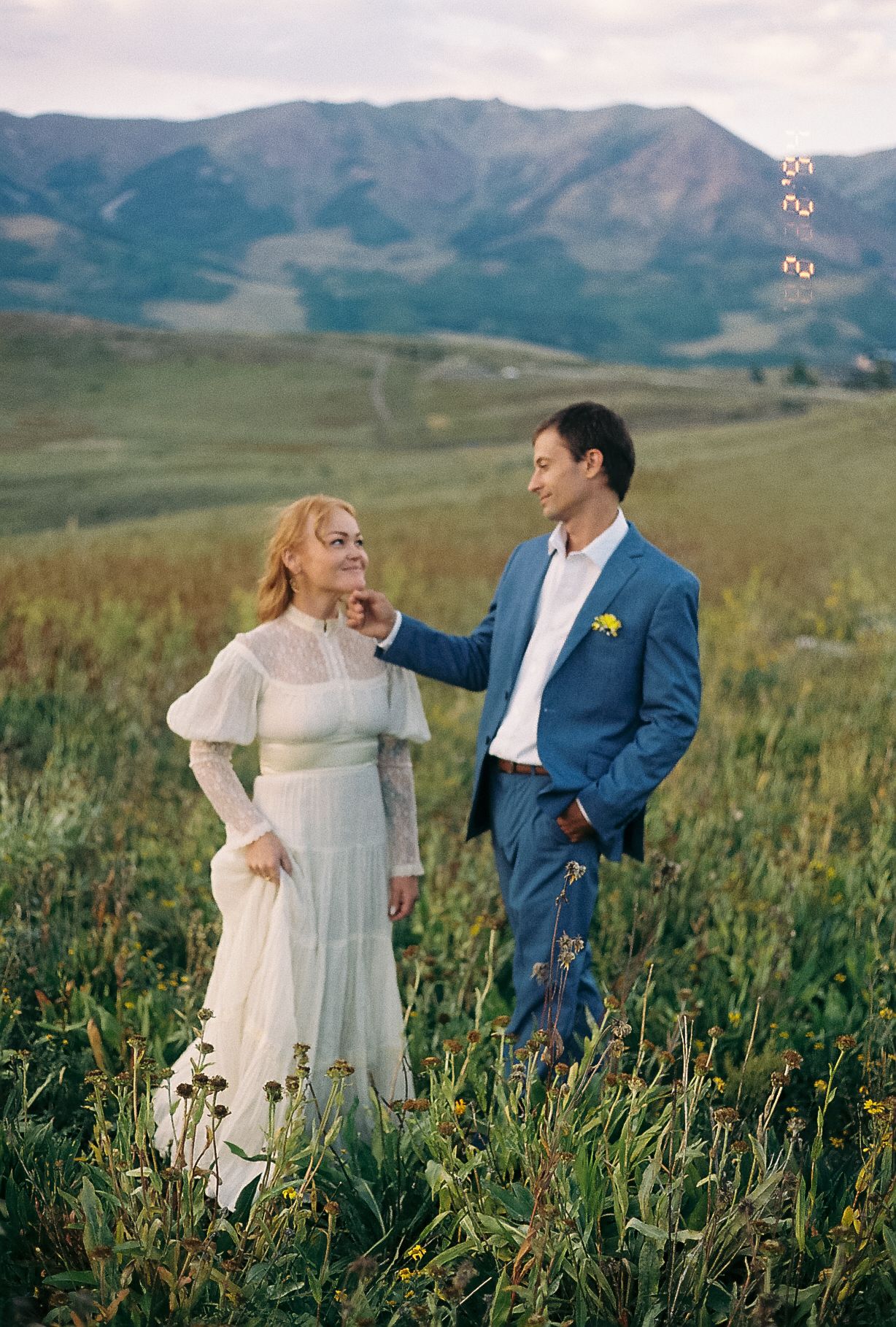 Groom in blue suit gently touches bride's face as she smiles in a wildflower meadow with mountain views.