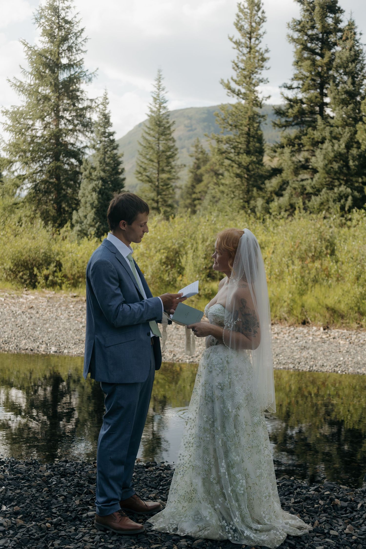 Couple exchanging vows outdoors by a mountain stream, groom in blue suit, bride in floral lace gown with veil.