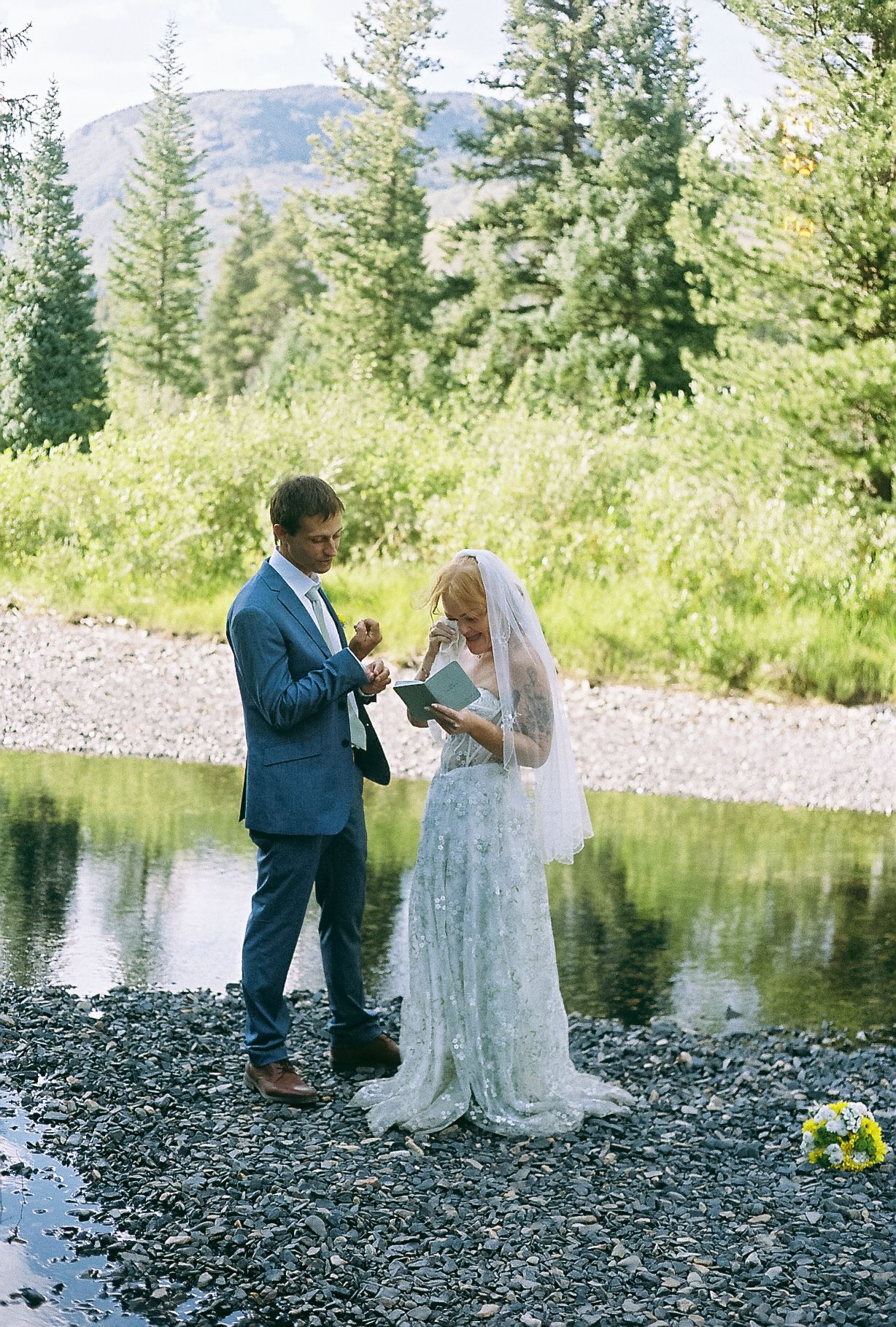 Bride reading vows to groom beside a rocky mountain stream with pine trees and peaks behind.