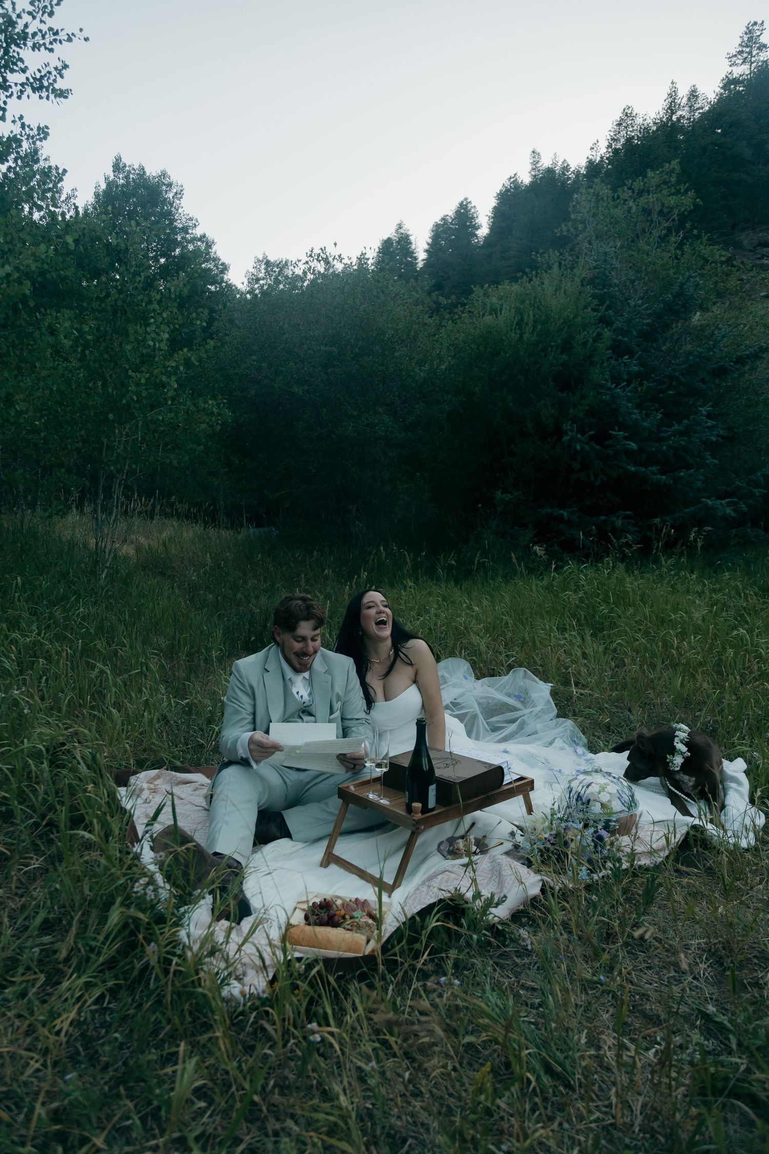 Laughing bride in white gown and groom in sage suit enjoying a picnic in a green forest meadow at dusk.