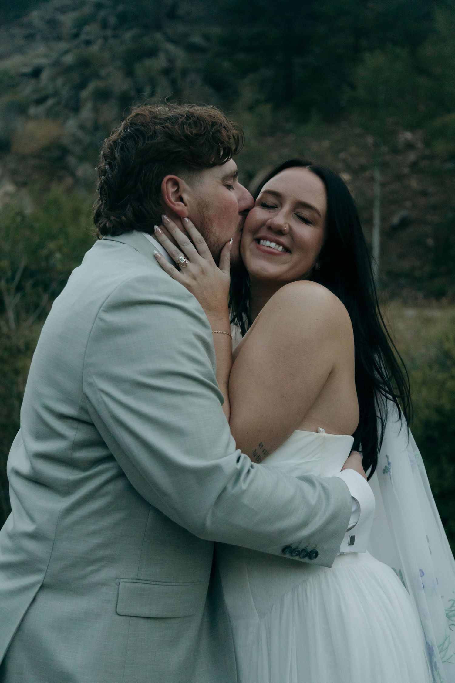 Groom kissing smiling bride on cheek with mountain backdrop during outdoor wedding portrait.