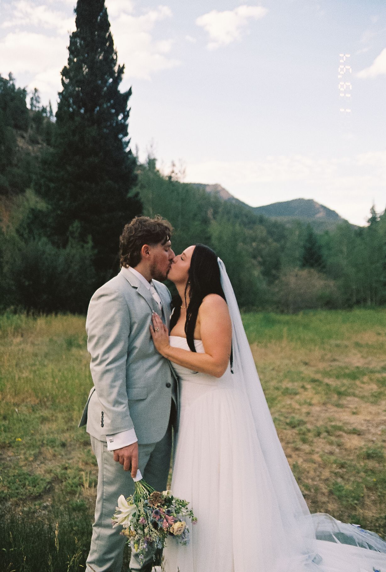 Bride and groom share first kiss in mountain meadow with wildflower bouquet on film photo.