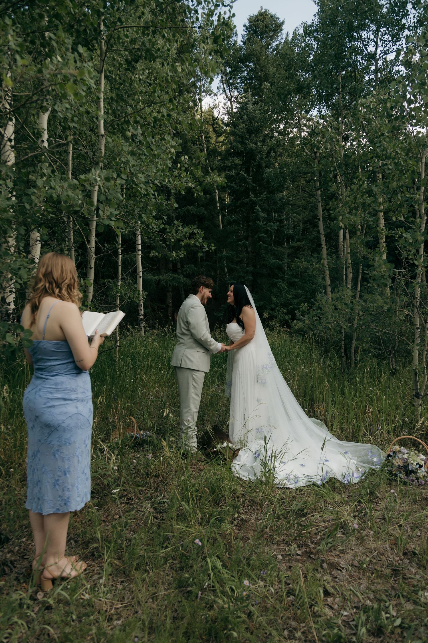 Bride and groom exchange vows in a forest clearing while an officiant reads from a book during an intimate outdoor wedding ceremony.