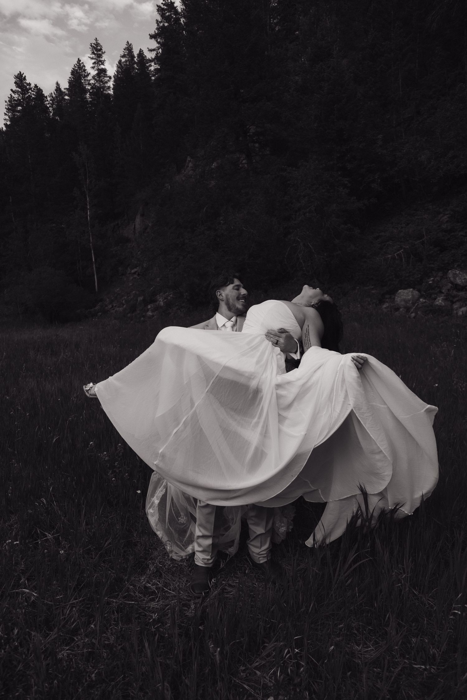 Black and white photo of bride and groom dancing in a field, her white dress flowing dramatically around them.