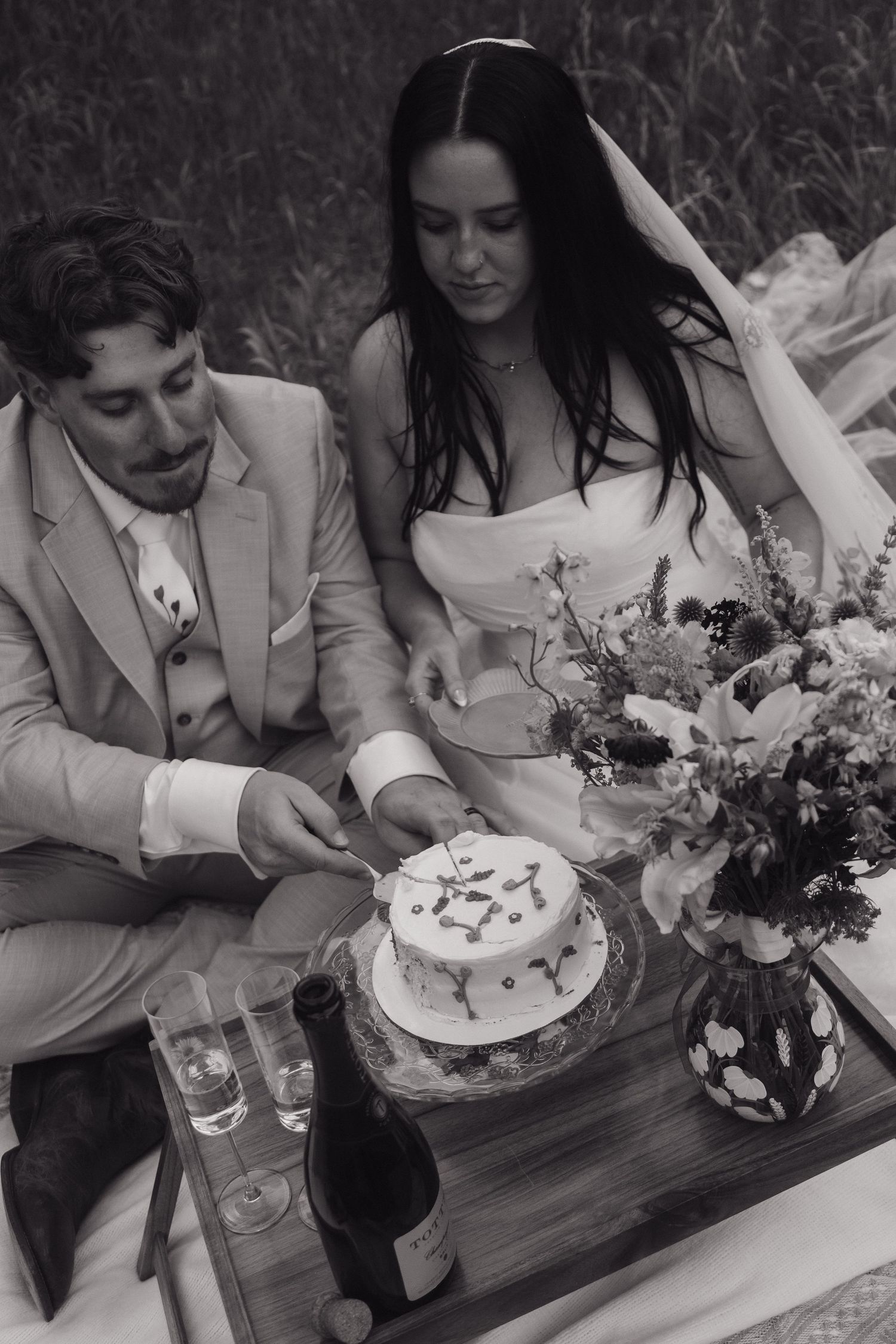 Black and white photo of bride and groom cutting a decorated wedding cake during an outdoor picnic celebration.