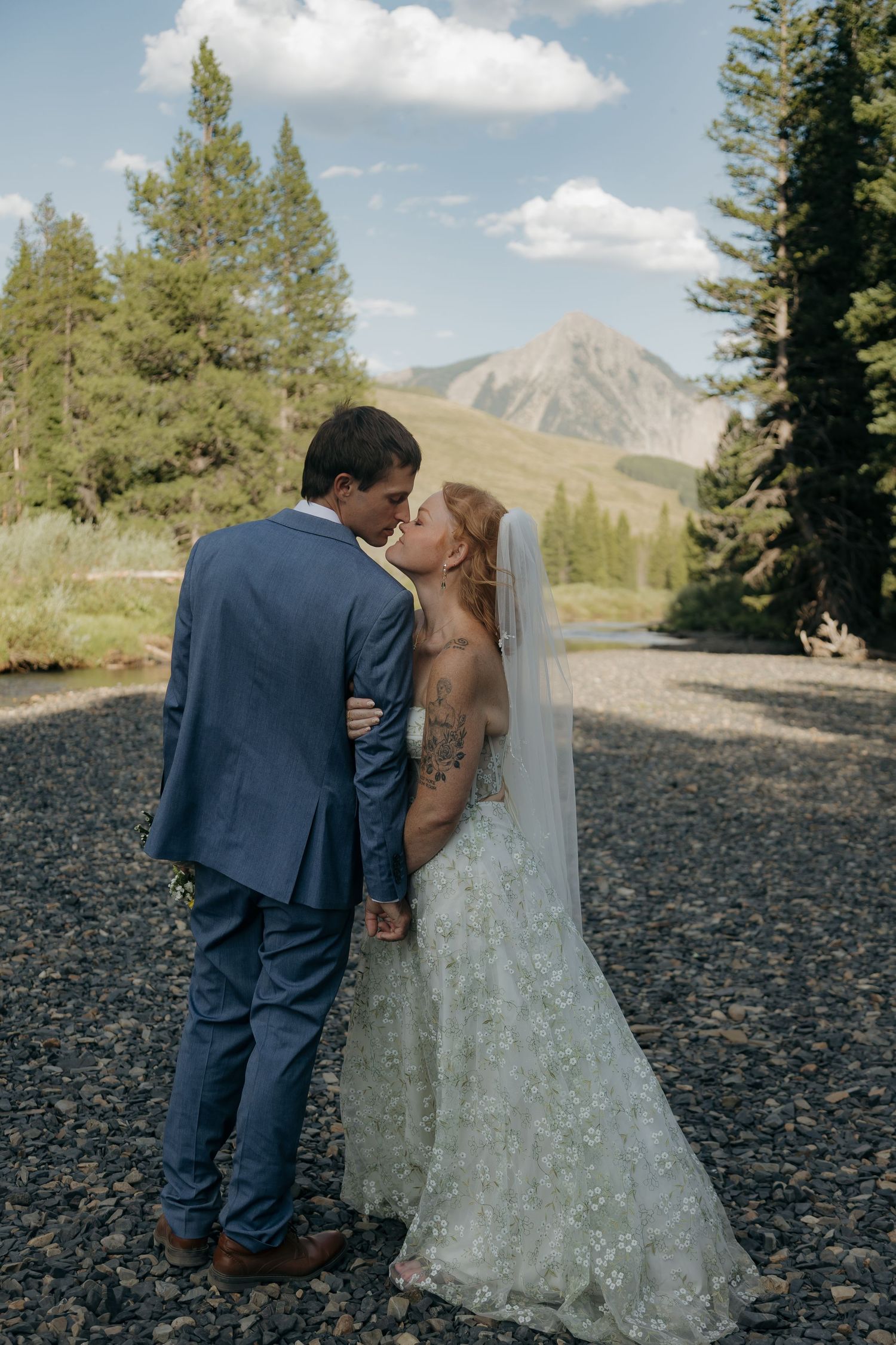 Bride and groom share a kiss beside a rocky mountain stream with a scenic peak in the background.