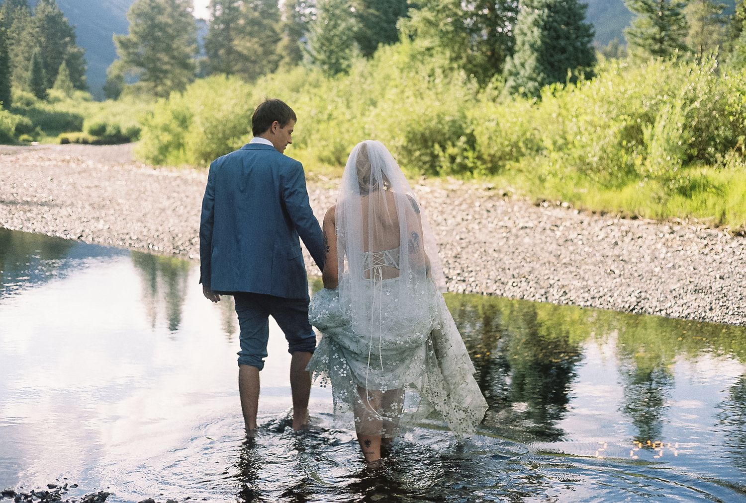 Bride and groom wade barefoot through shallow mountain stream holding hands in wedding attire.