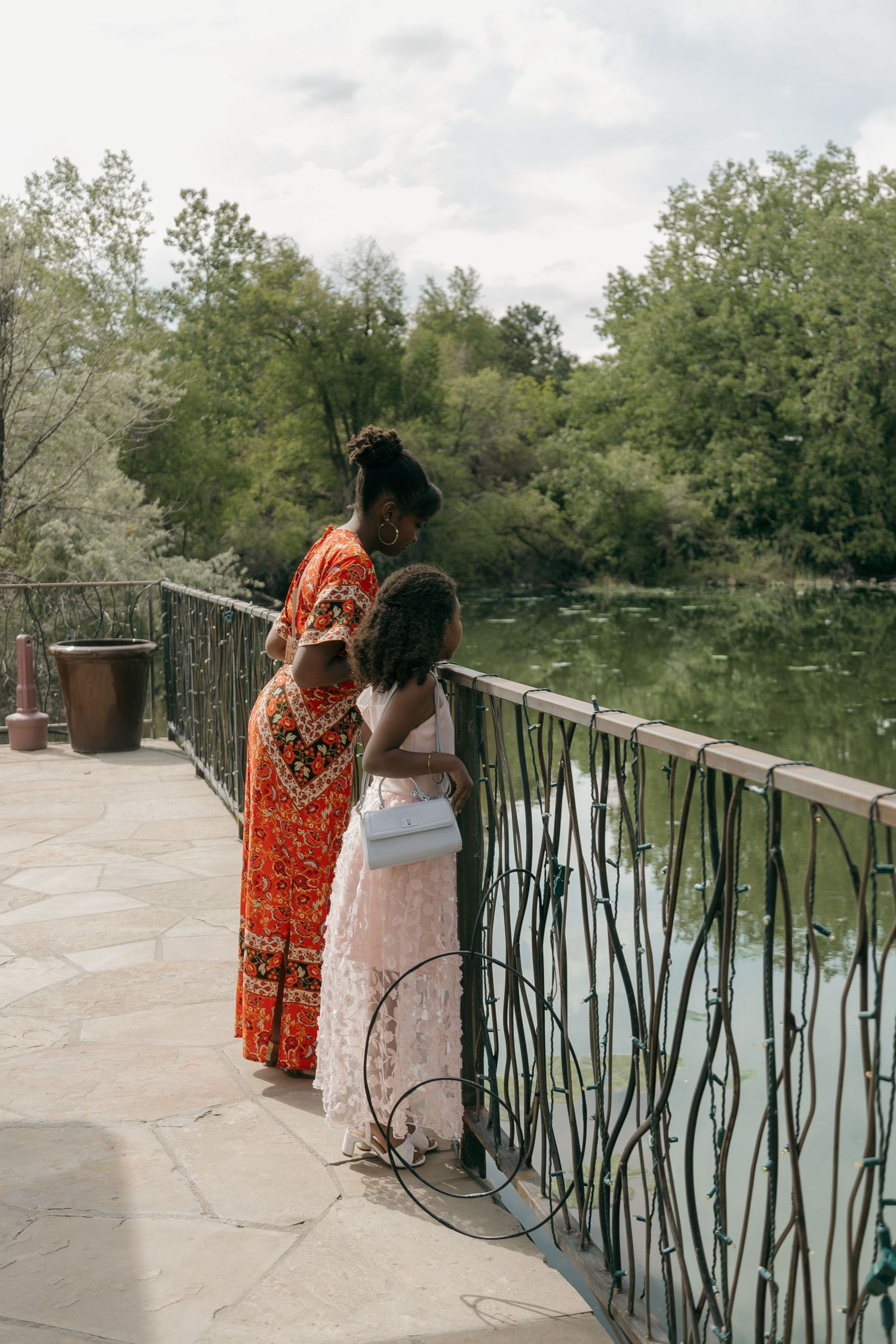 Mother and daughter in colorful attire standing on a scenic bridge overlooking a tranquil pond.
