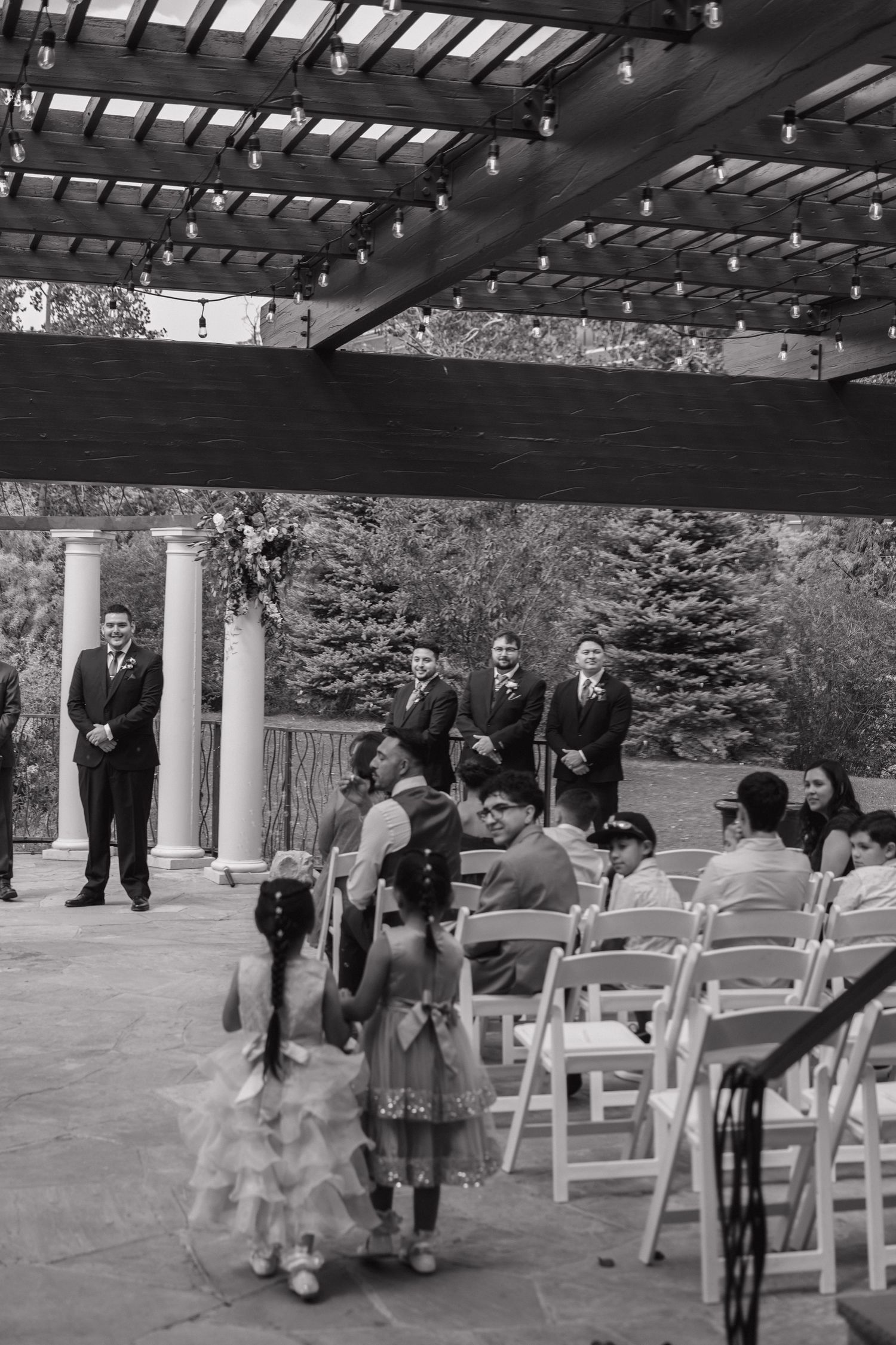Wedding ceremony with flower girls walking the aisle under a pergola with string lights and white columns.