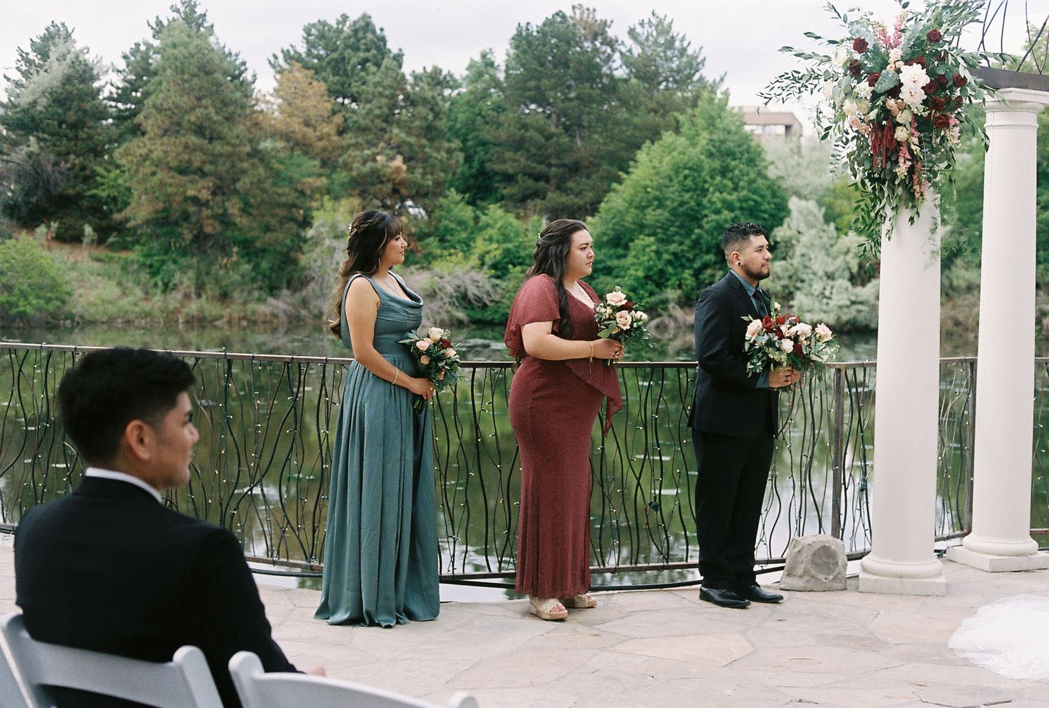 Bridesmaids in teal and burgundy dresses holding bouquets standing at outdoor waterfront wedding ceremony.